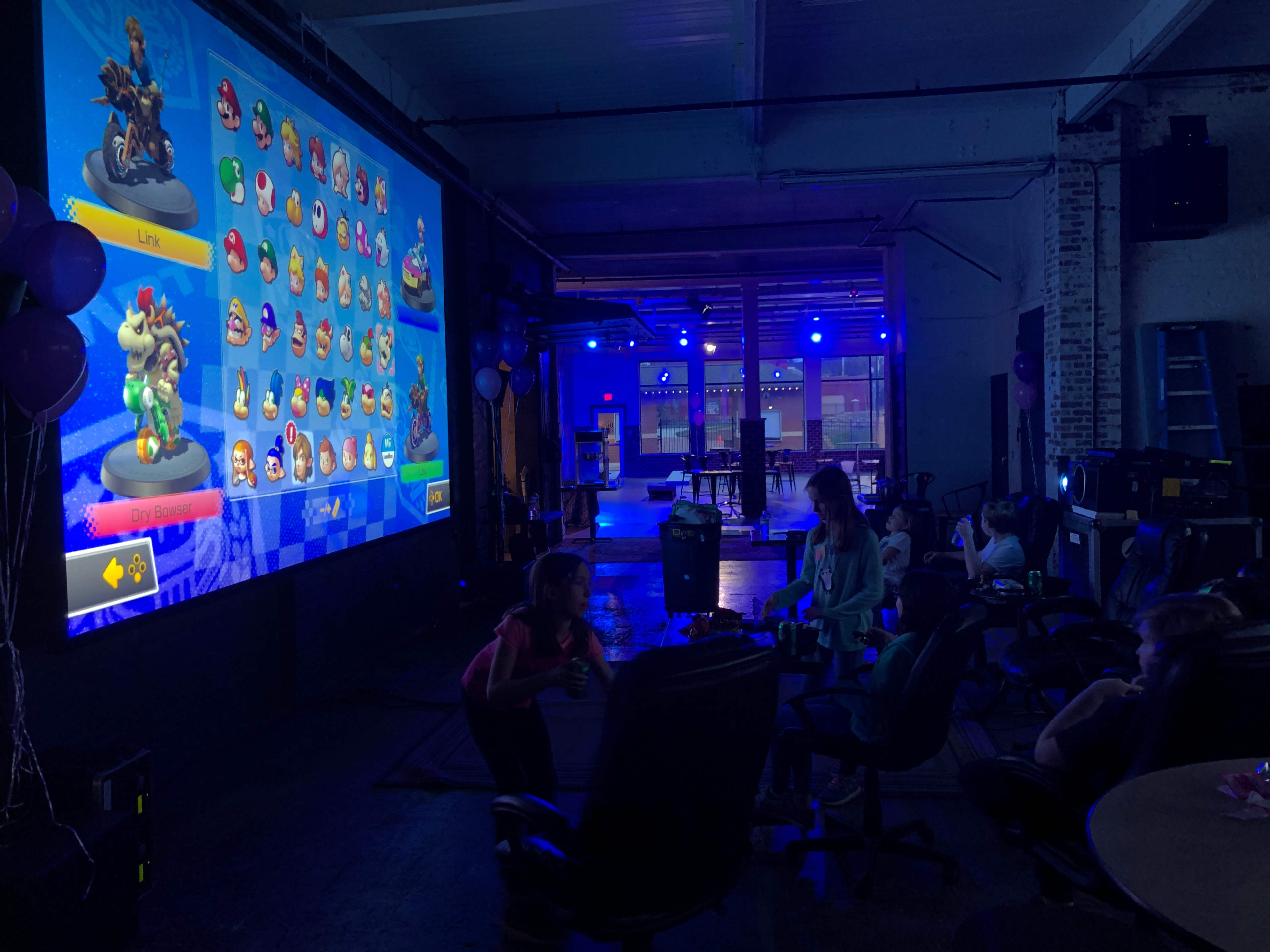 The scene shows a gaming event in a spacious room with a large screen displaying characters from a racing game, while children sit in gaming chairs in front of it.
