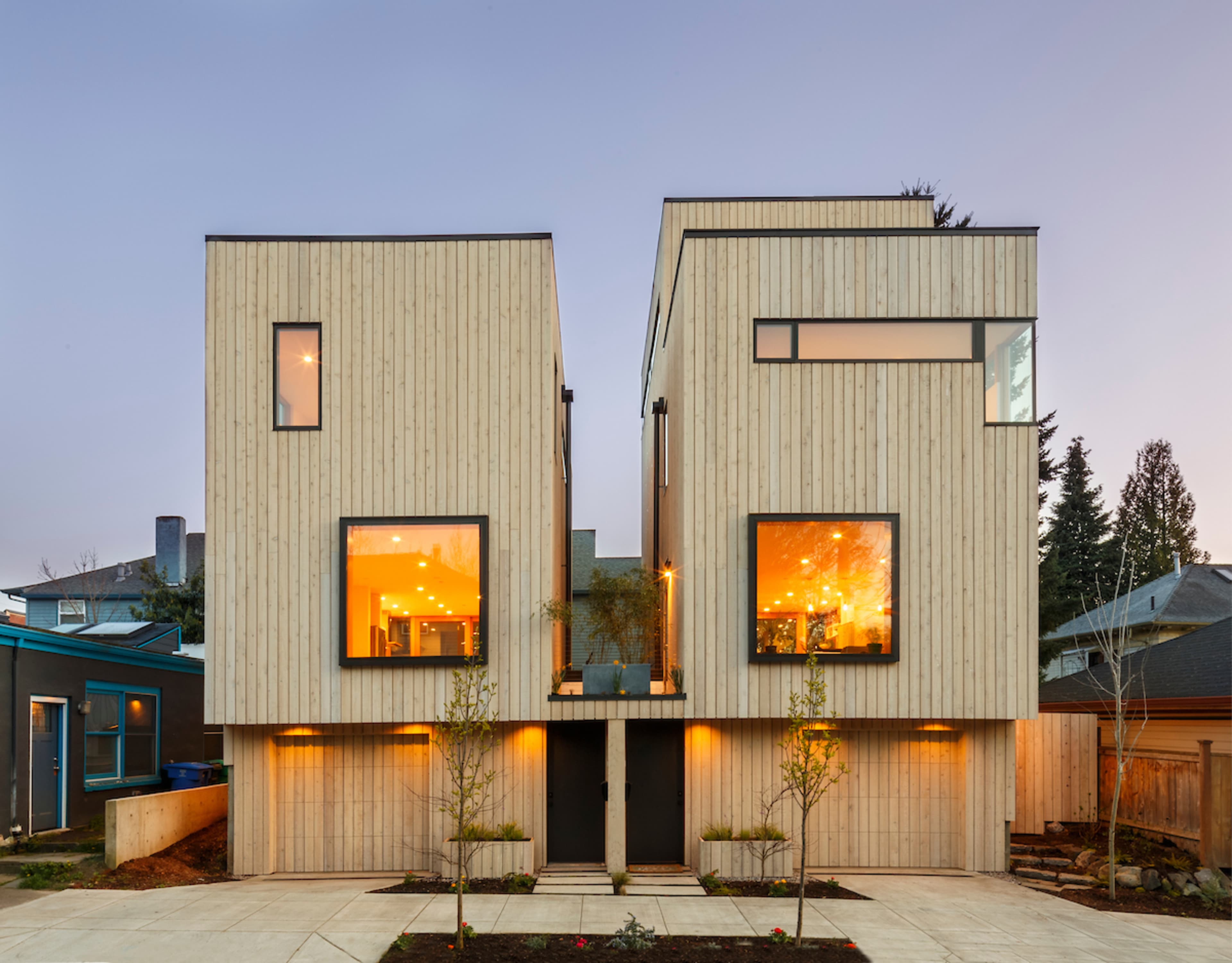 A modern, two-story wooden house with large windows, illuminated interiors, and a symmetrical facade.