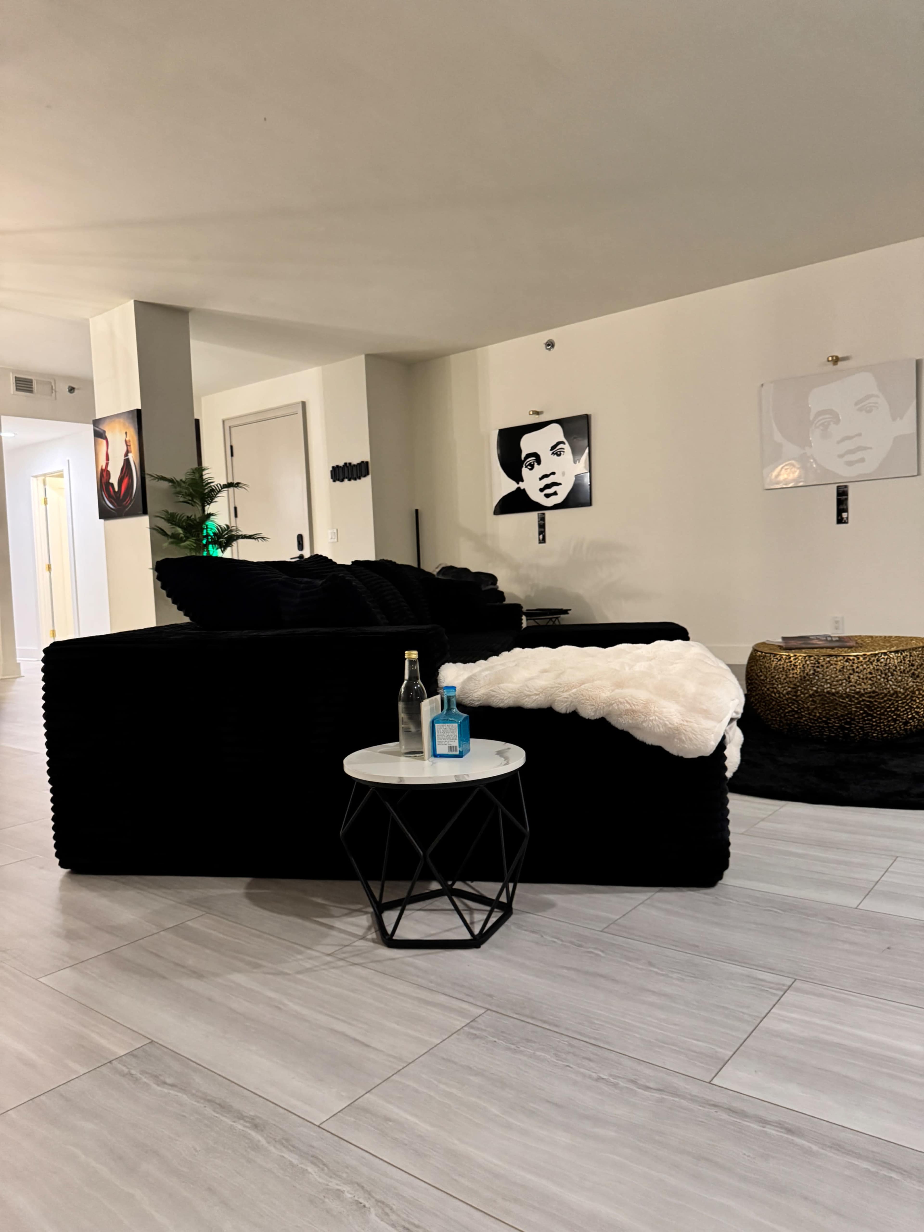 The image shows a modern living room with a black couch, a small round table featuring a bottle and a blue object, and artwork of faces on the walls.