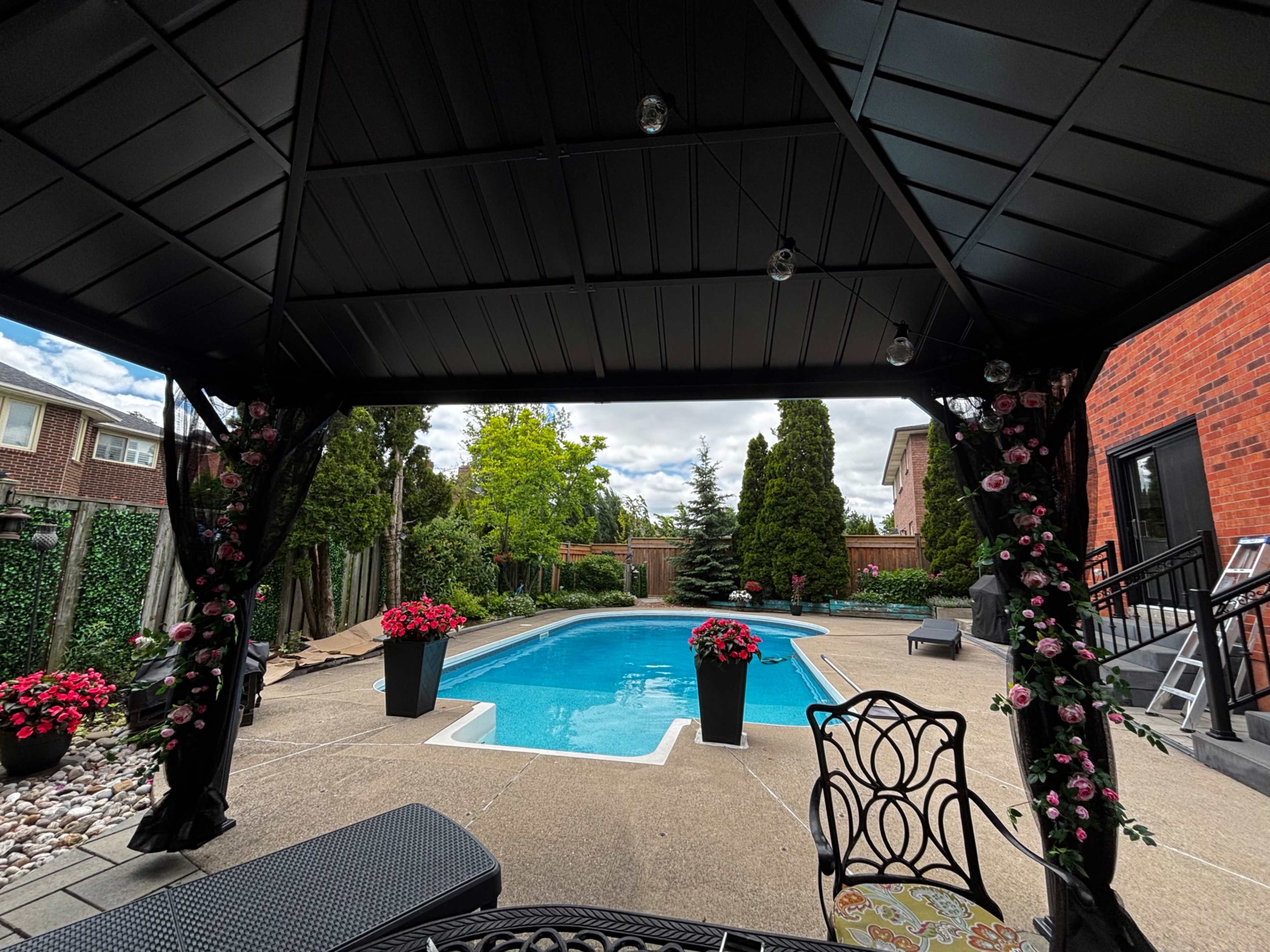 The image shows a backyard pool area with a gazebo, surrounded by potted flowers and lush greenery.