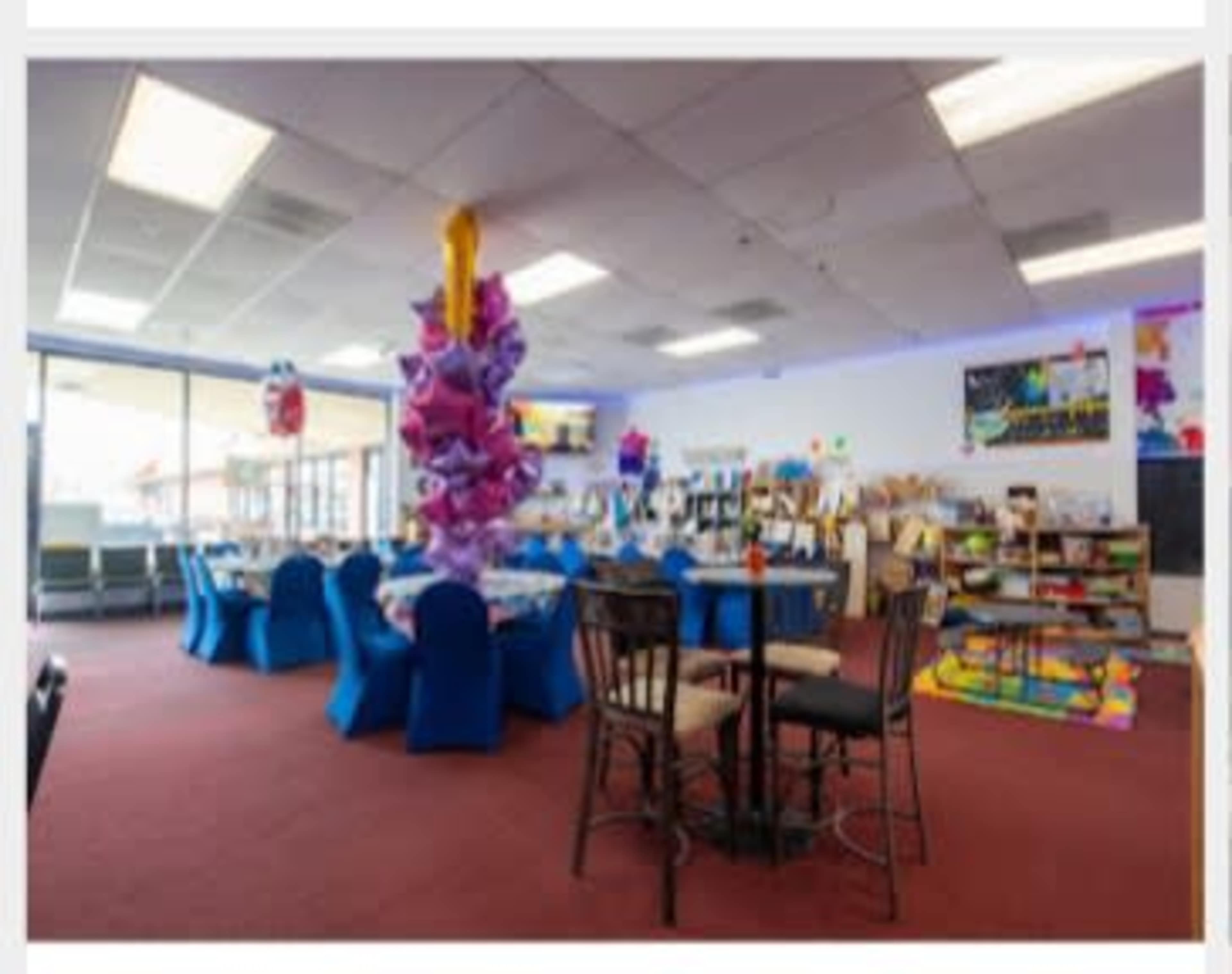 A decorated event space with blue table covers, a central colorful decoration hanging from the ceiling, and various activity stations visible along the walls.