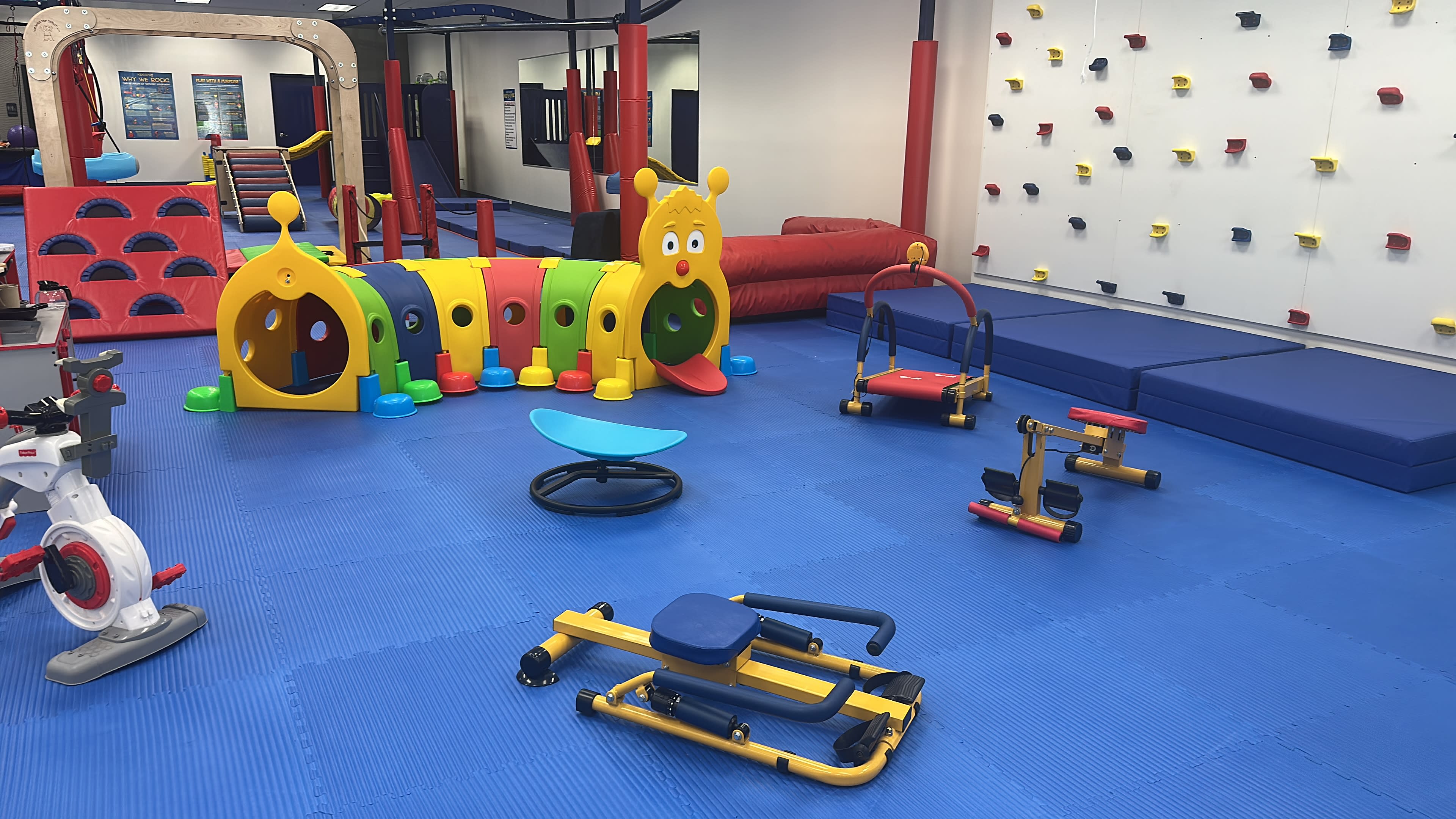The image shows a brightly colorful indoor play area featuring a variety of children's play equipment, including a caterpillar climbing structure, balance toys, and soft mats.