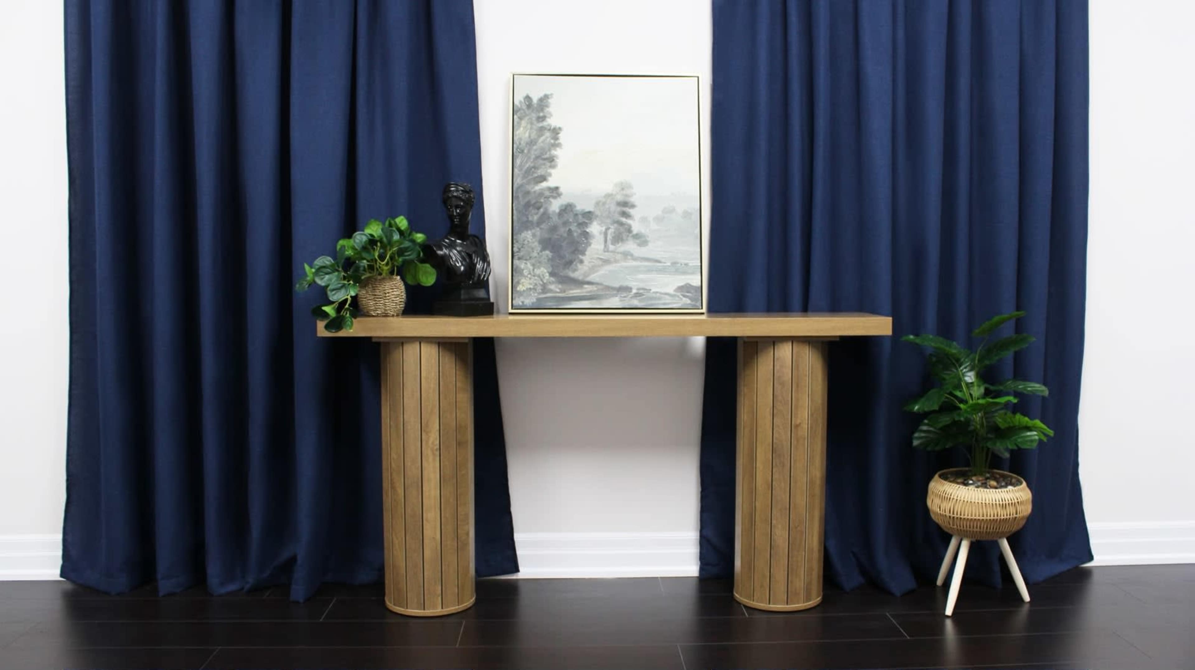 A wooden console table with two cylindrical legs holds a framed artwork, accompanied by a bust and two potted plants, set against navy curtains.