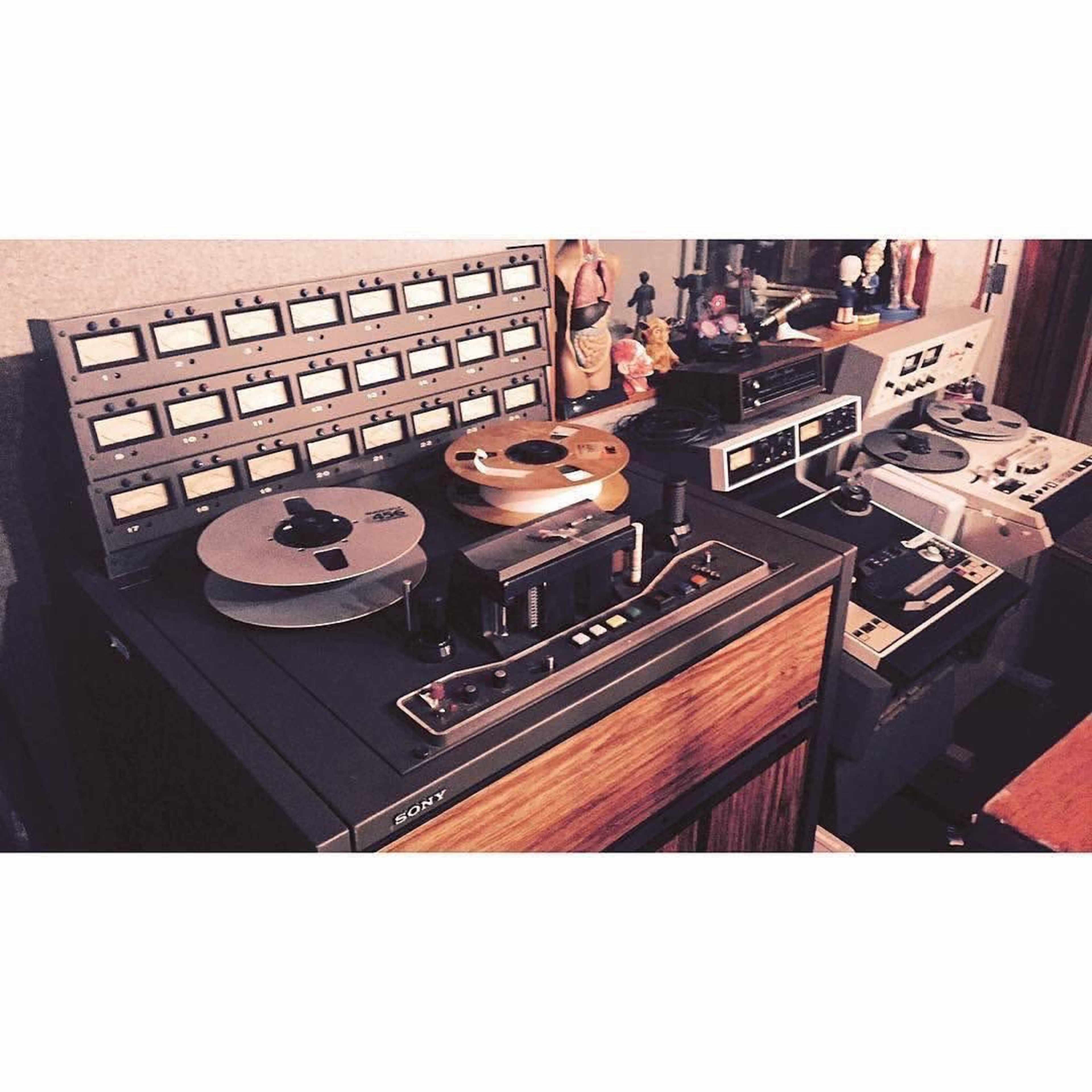 A vintage audio setup featuring multiple reel-to-reel tape recorders and a large control panel with numerous buttons and dials.