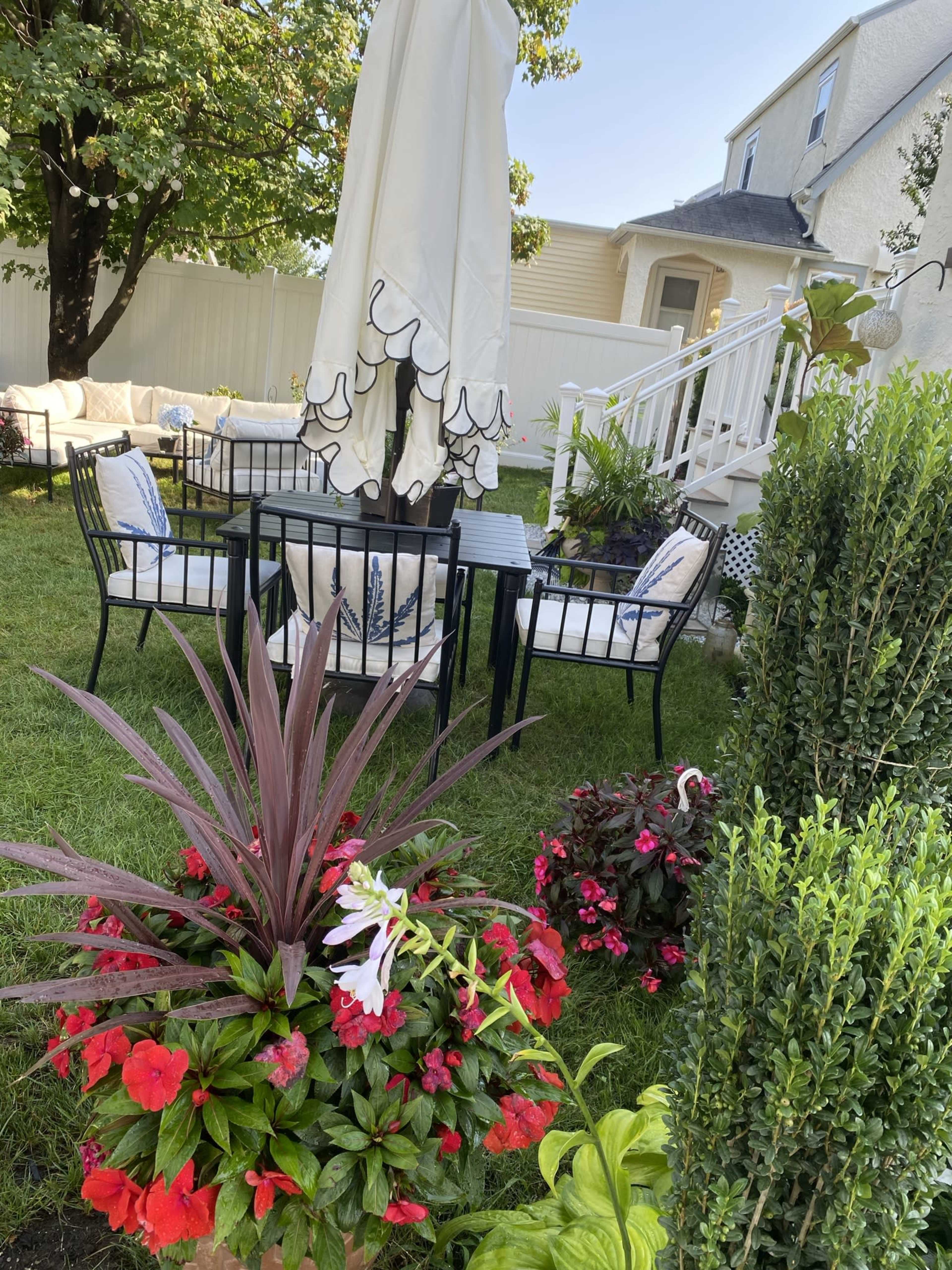 A backyard patio area with a table and chairs beneath a large umbrella, surrounded by various flowering plants.