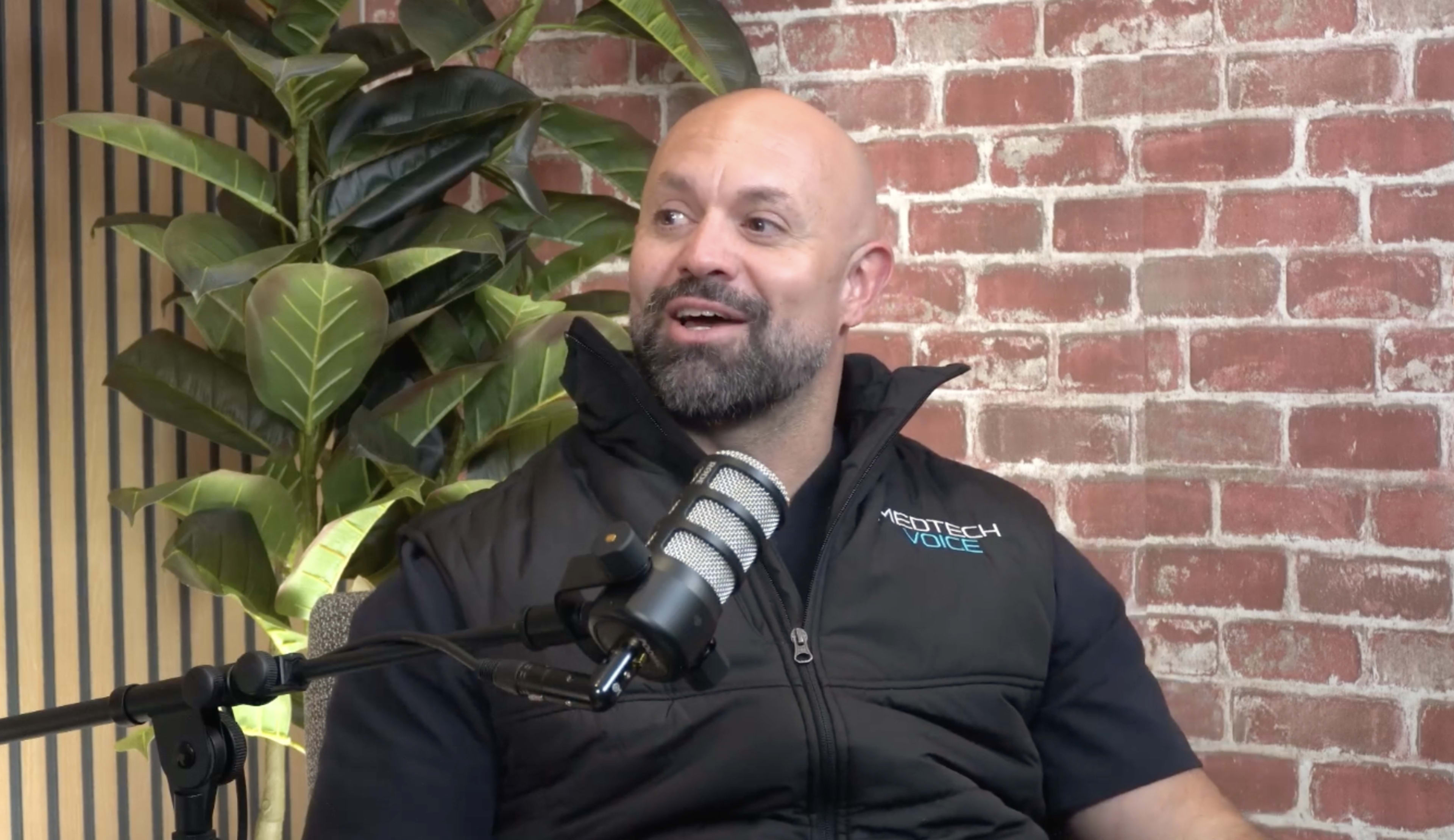 A man with a shaved head and beard is seated in front of a microphone, smiling, with a brick wall and a green plant in the background.