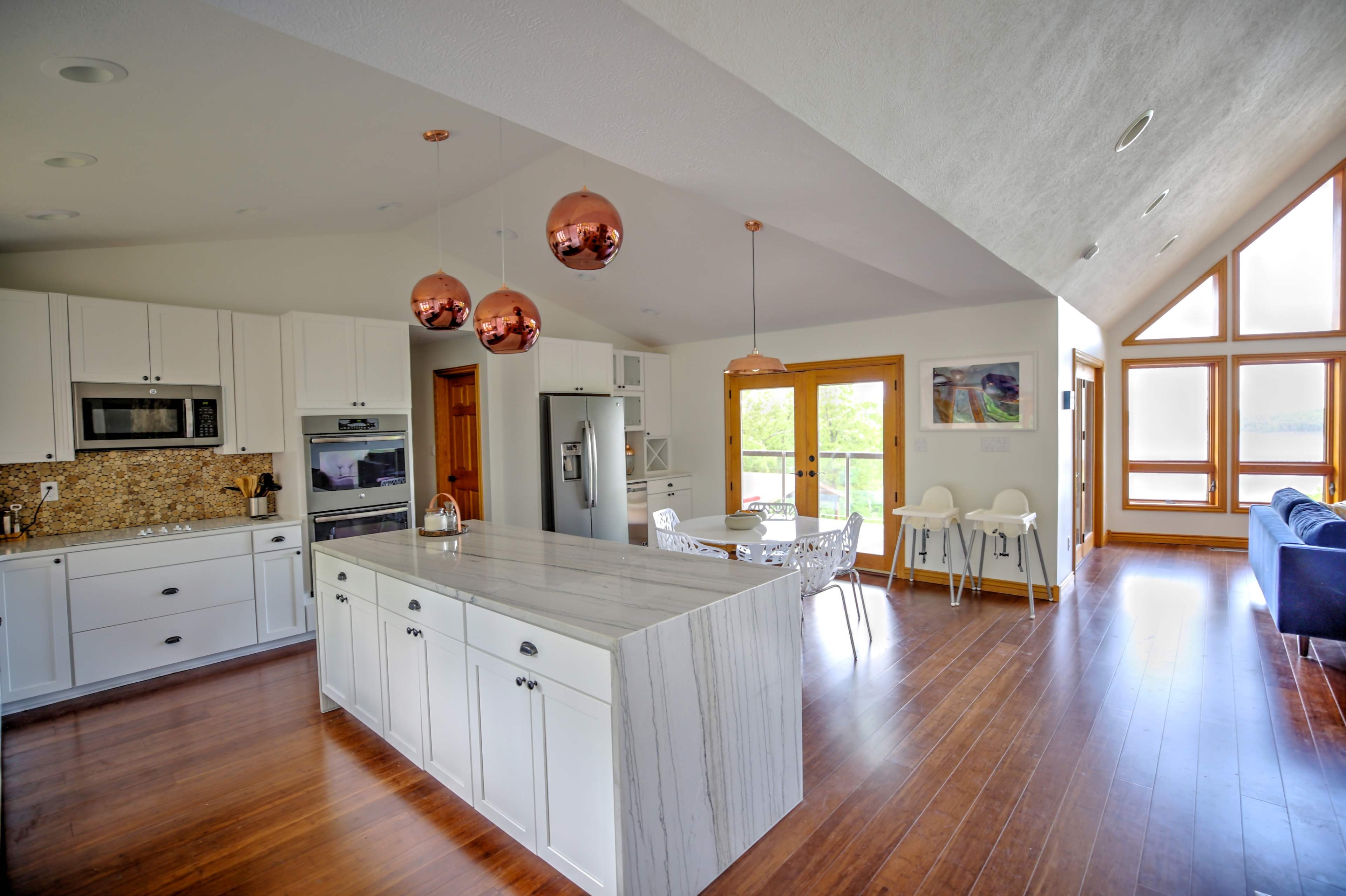 A modern kitchen with white cabinets, a marble island, and copper pendant lights, adjacent to a dining area with a table and chairs under large windows.