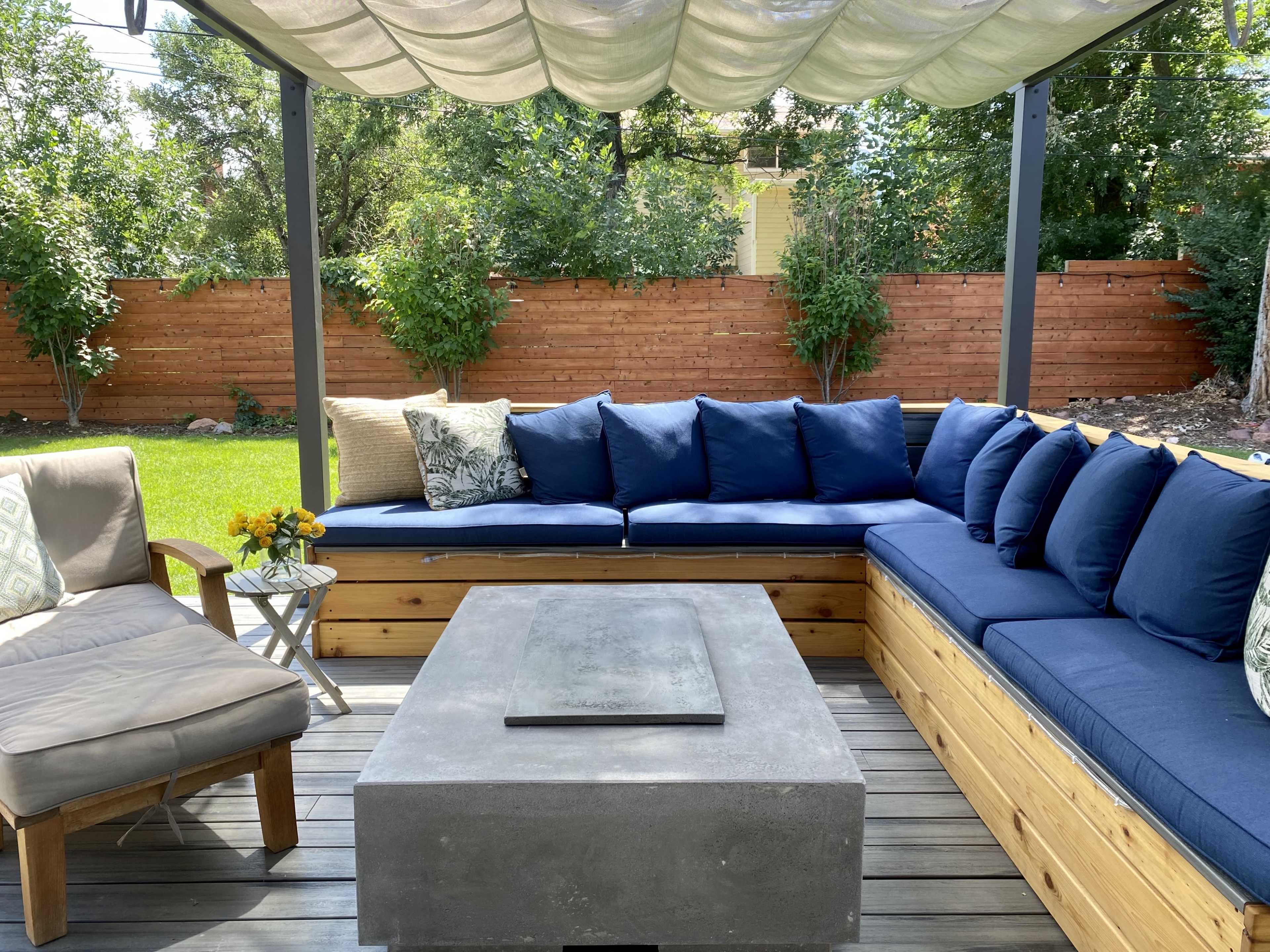 A outdoor seating area features a large sectional couch with blue cushions, a concrete coffee table, and a small side table, all under a fabric canopy.