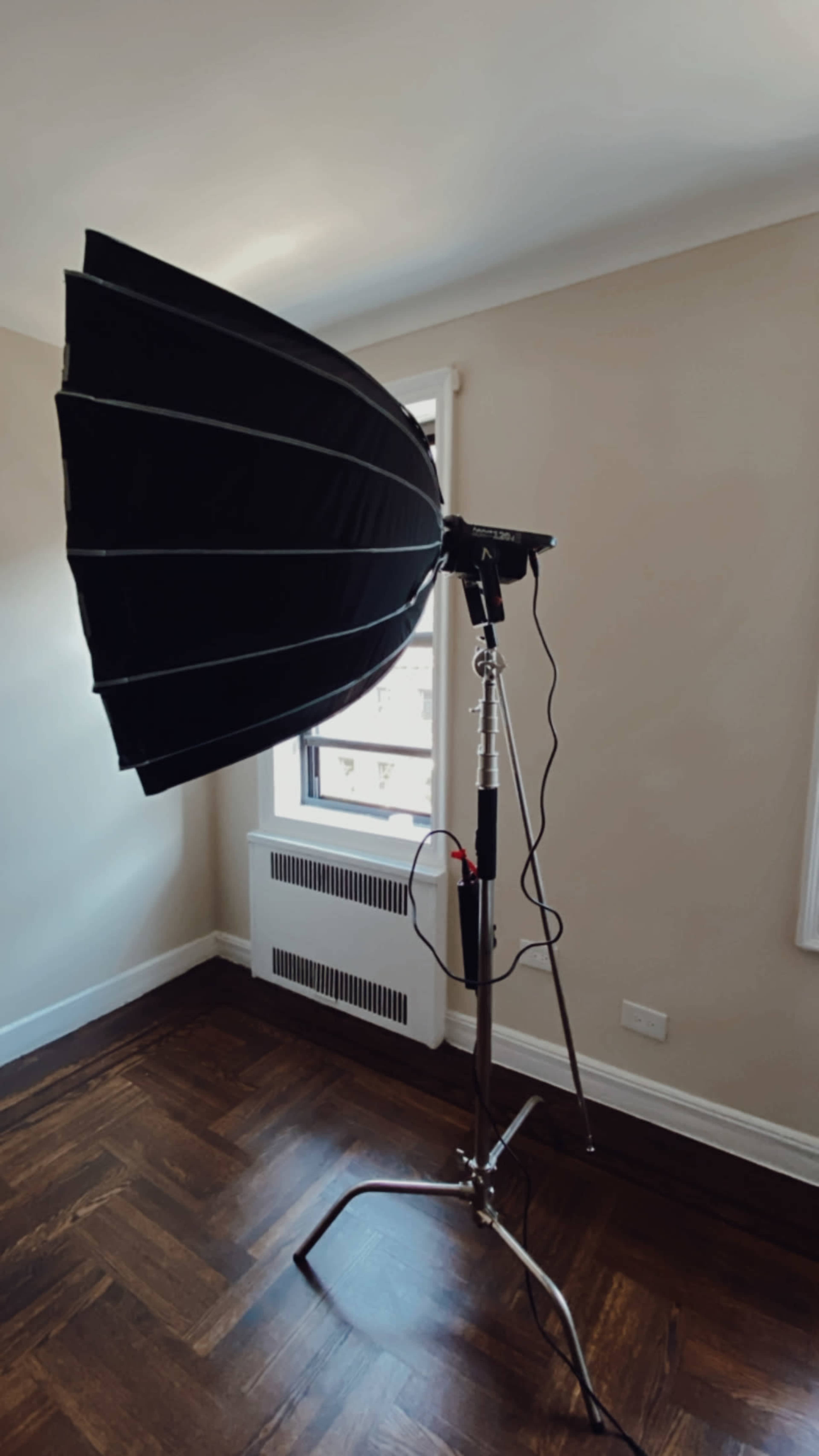 A large softbox light is set up in a sparsely furnished room with hardwood floors and a window.