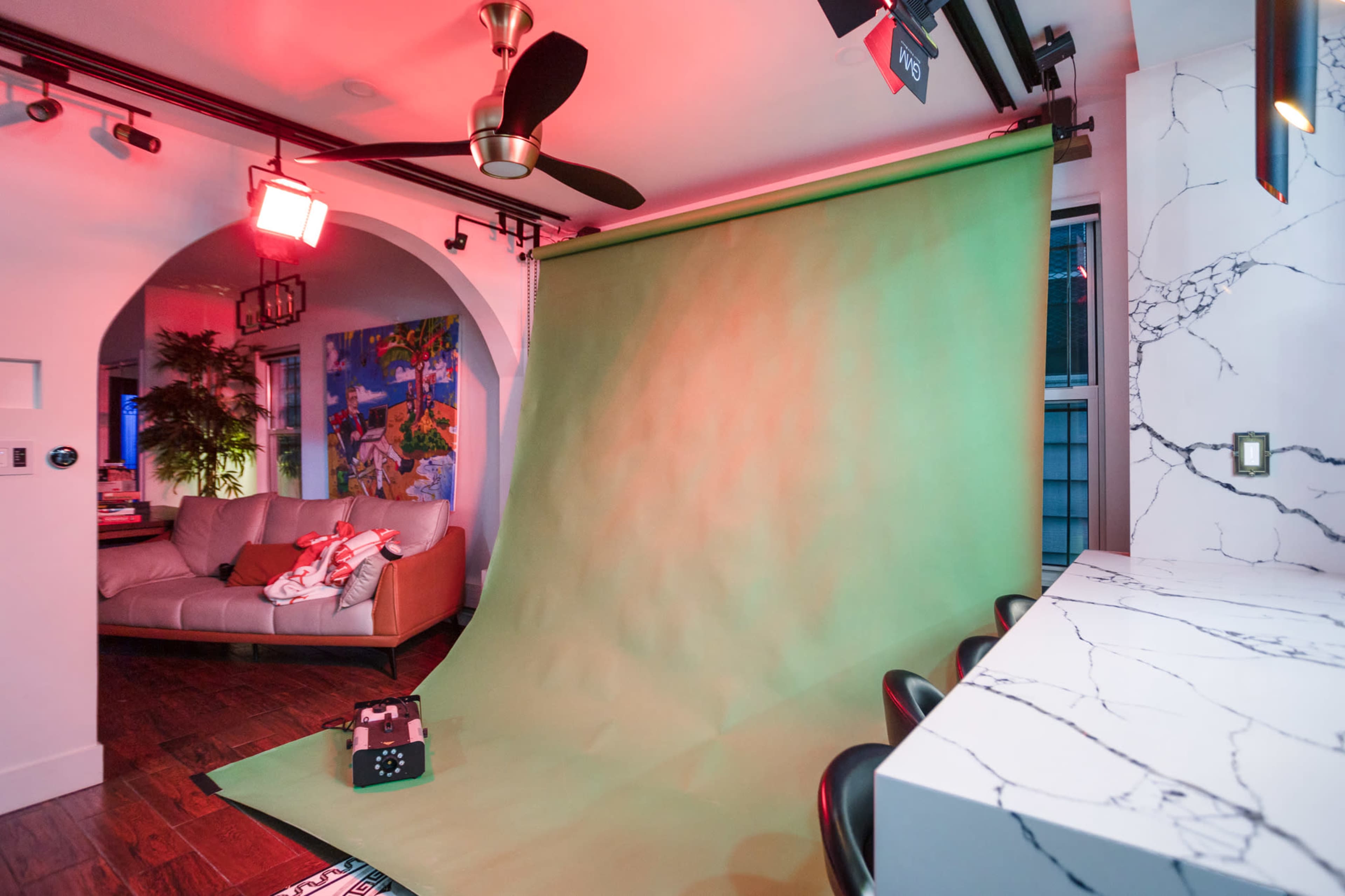 A room with a green backdrop set up for photography, accompanied by a couch, decorative artwork, and modern kitchen elements.
