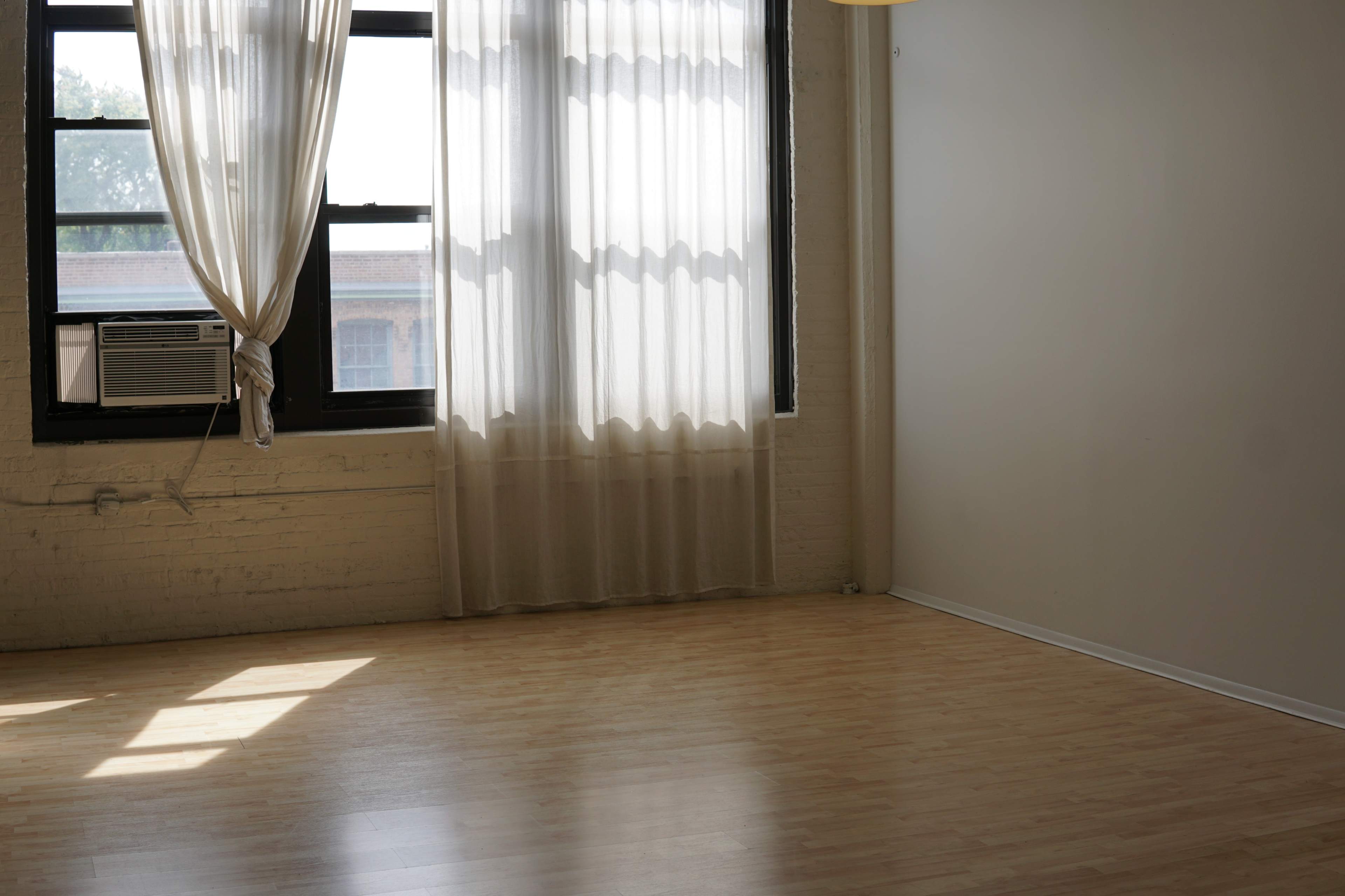 A bright room with a wooden floor, a large window with sheer curtains, and an air conditioning unit mounted near the window.