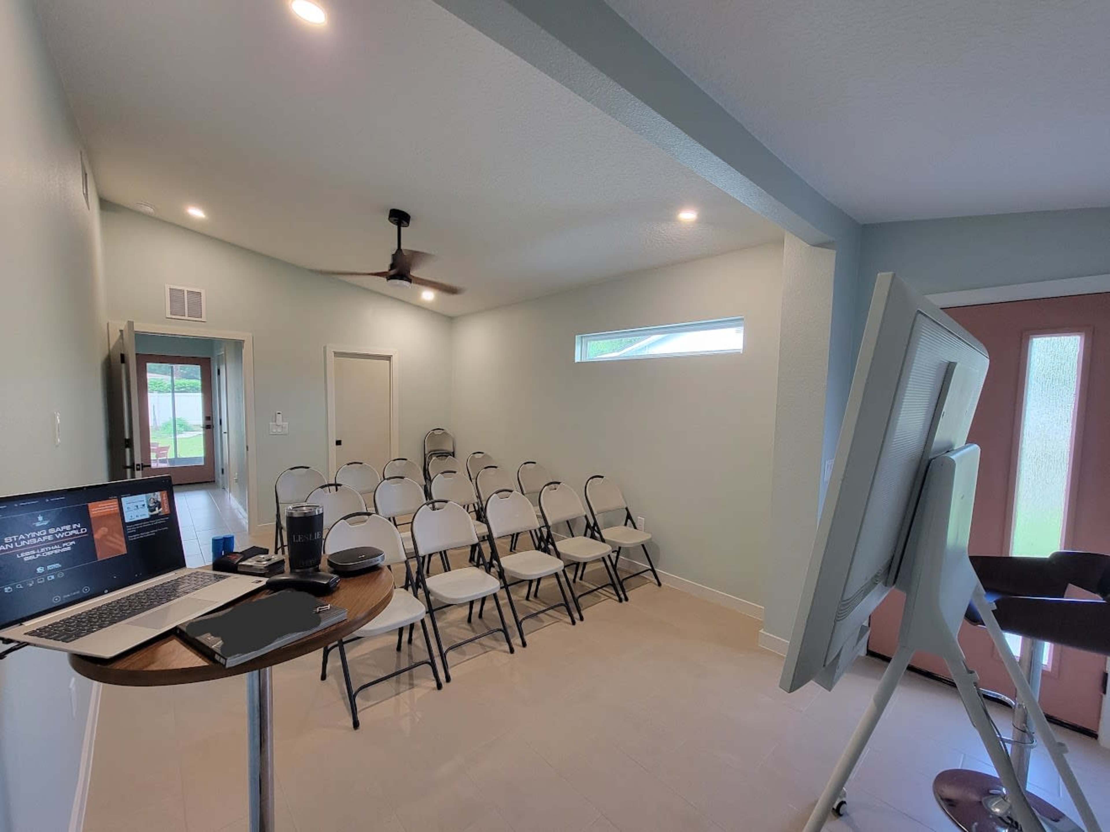 A small, lightly furnished room features rows of white chairs facing a laptop and a standing screen near a door with pink accents.