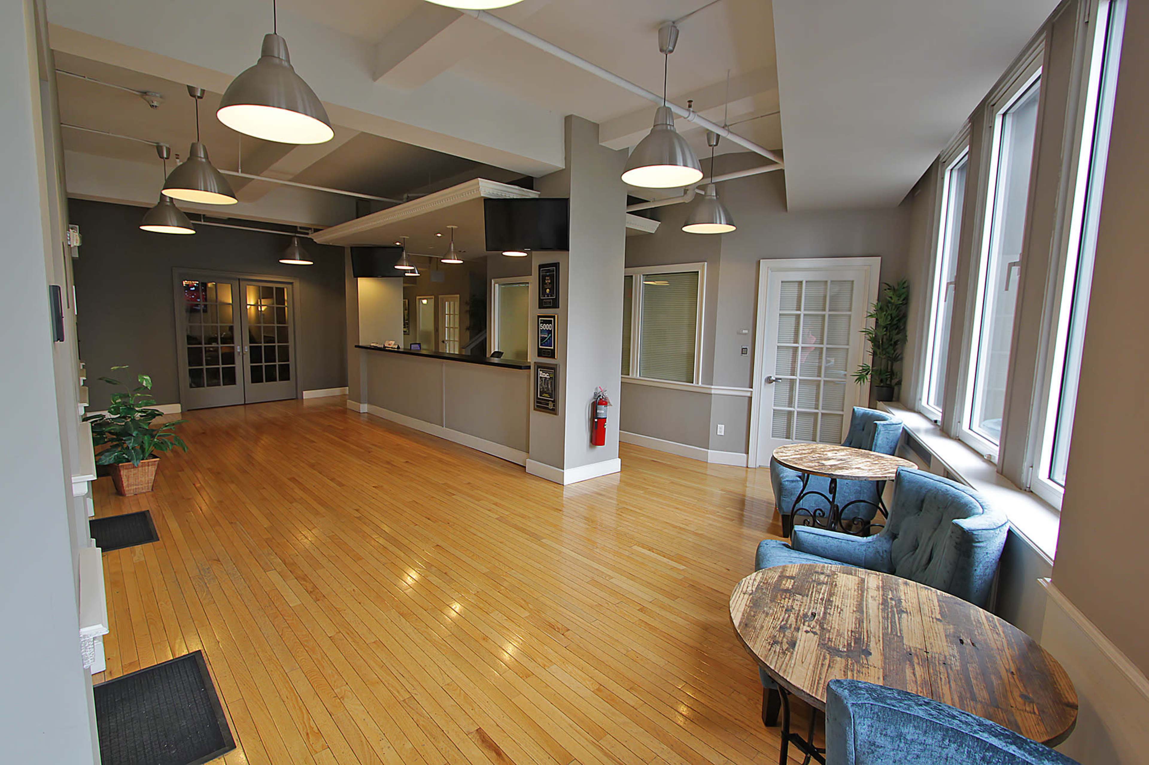 The image shows a spacious, well-lit reception area with wooden floors, blue upholstered chairs, small round tables, and a front desk.