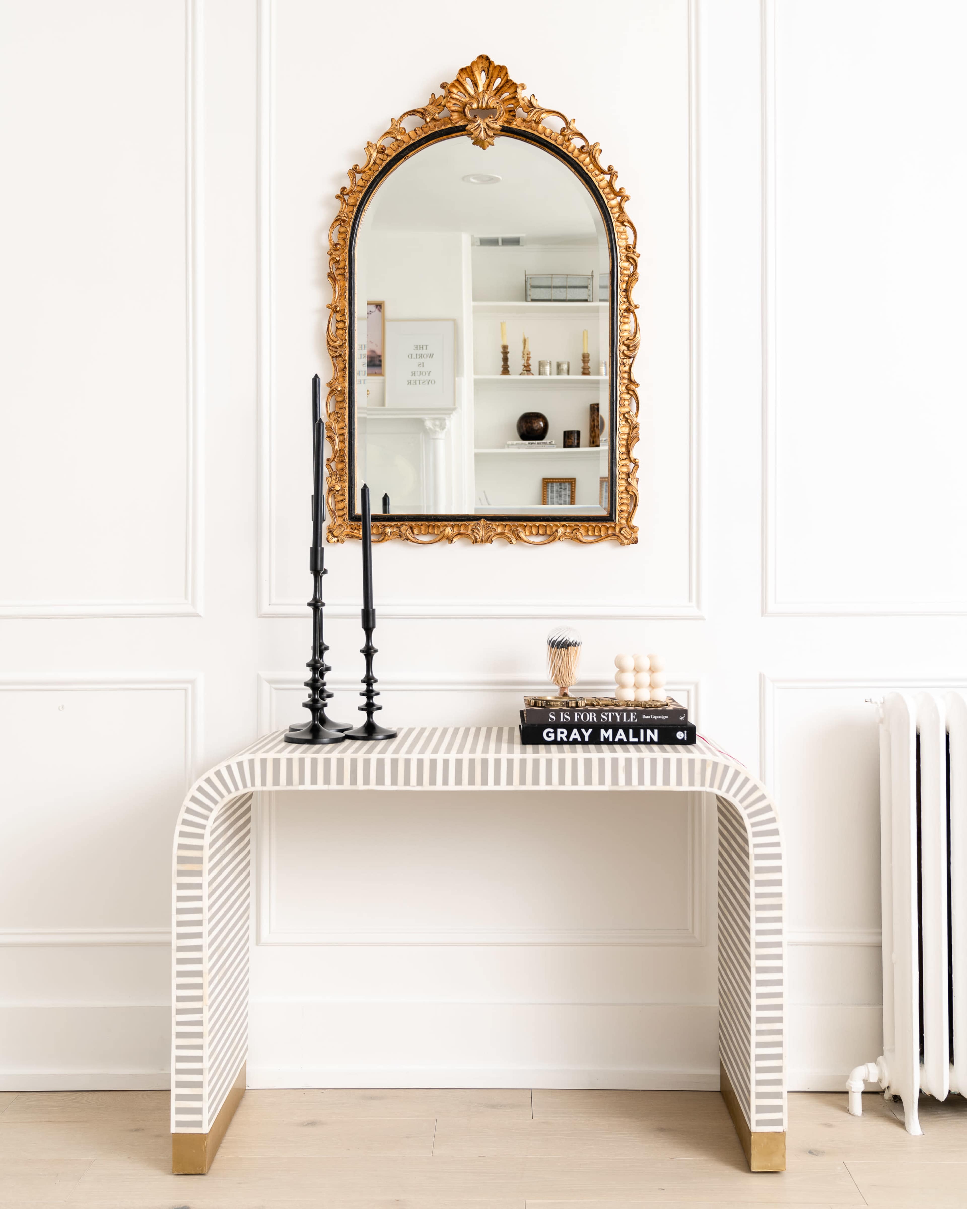 A striped console table with a gold-framed mirror and black candlesticks is positioned against a white wall.
