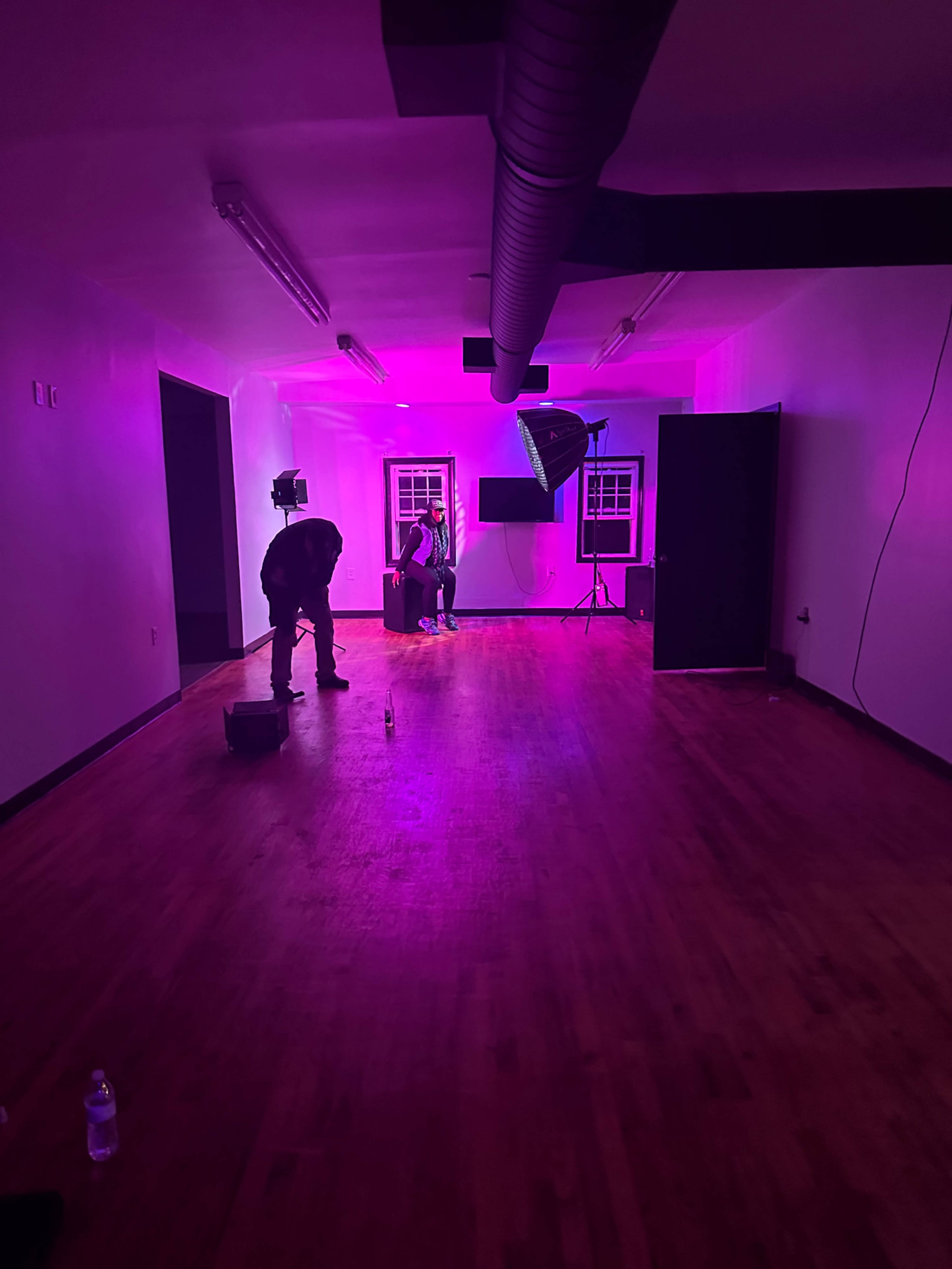 A photographer sets up lighting equipment in a sparsely furnished room with purple illumination while a subject sits on a stool.