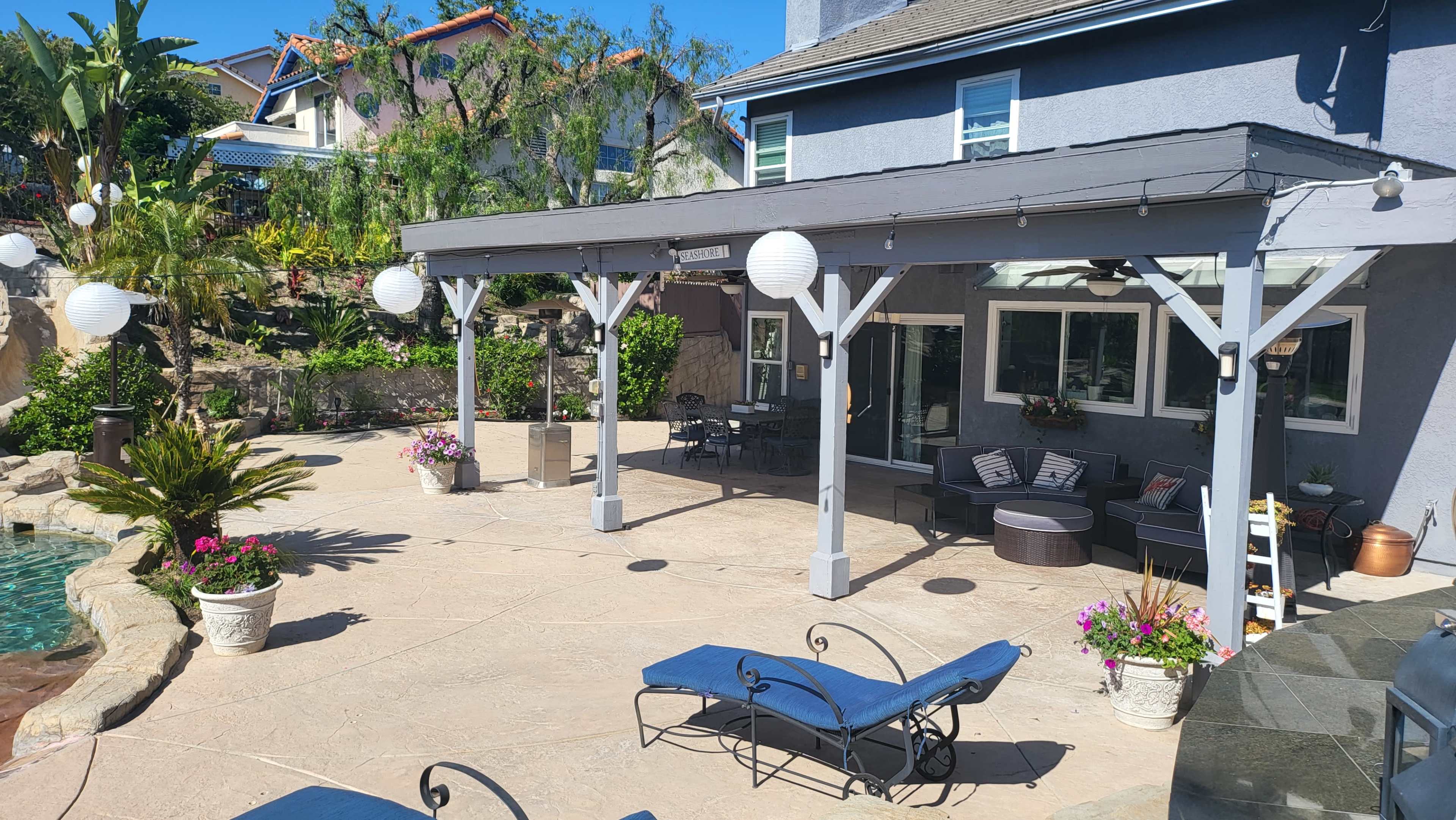 A spacious outdoor patio area with lounge chairs, potted plants, and decorative lanterns under a shaded structure adjacent to a pool and landscaped yard.