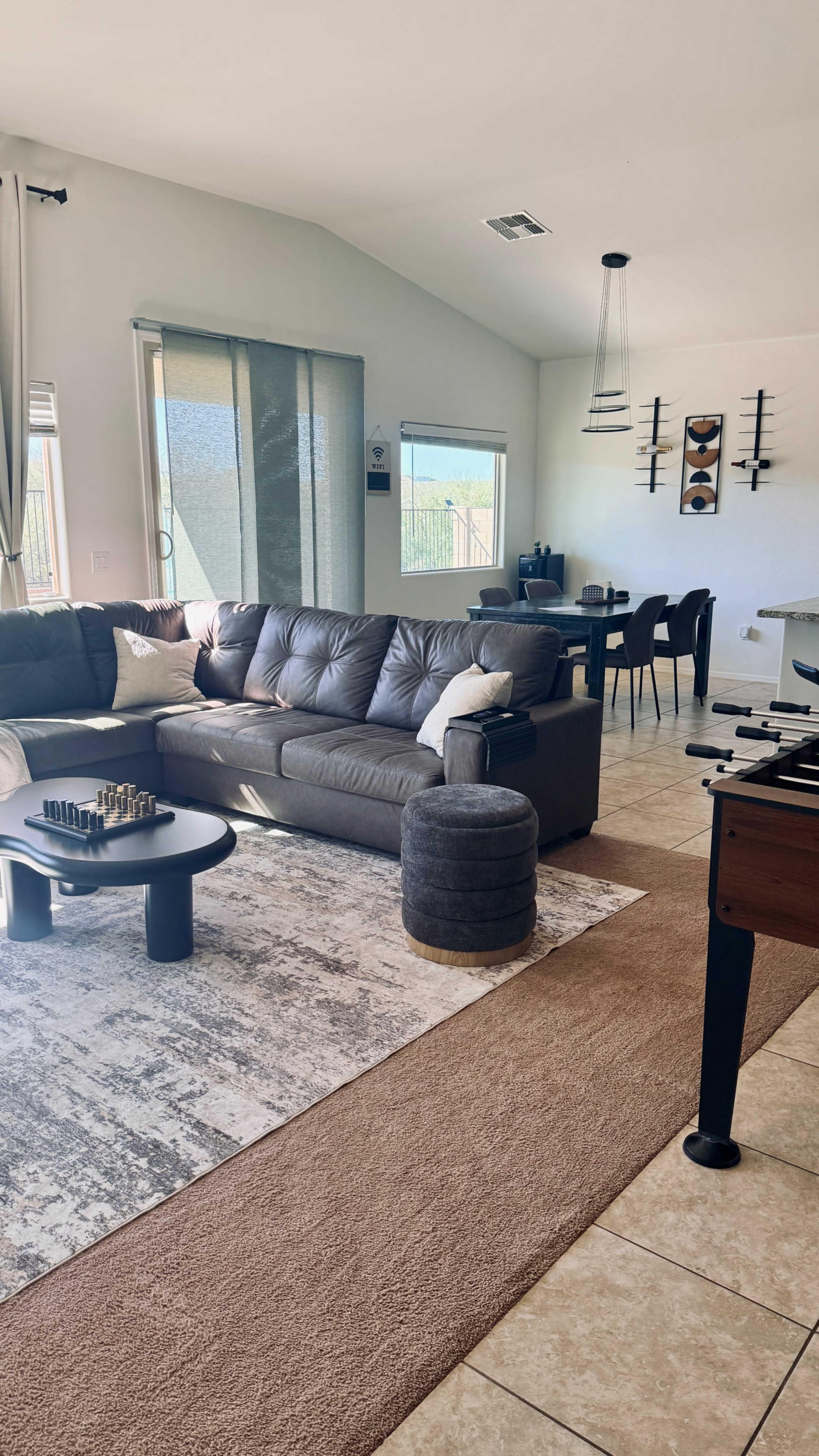 The image shows a modern living room with a gray sectional sofa, a round coffee table, a game table, and a dining area in an open layout space.