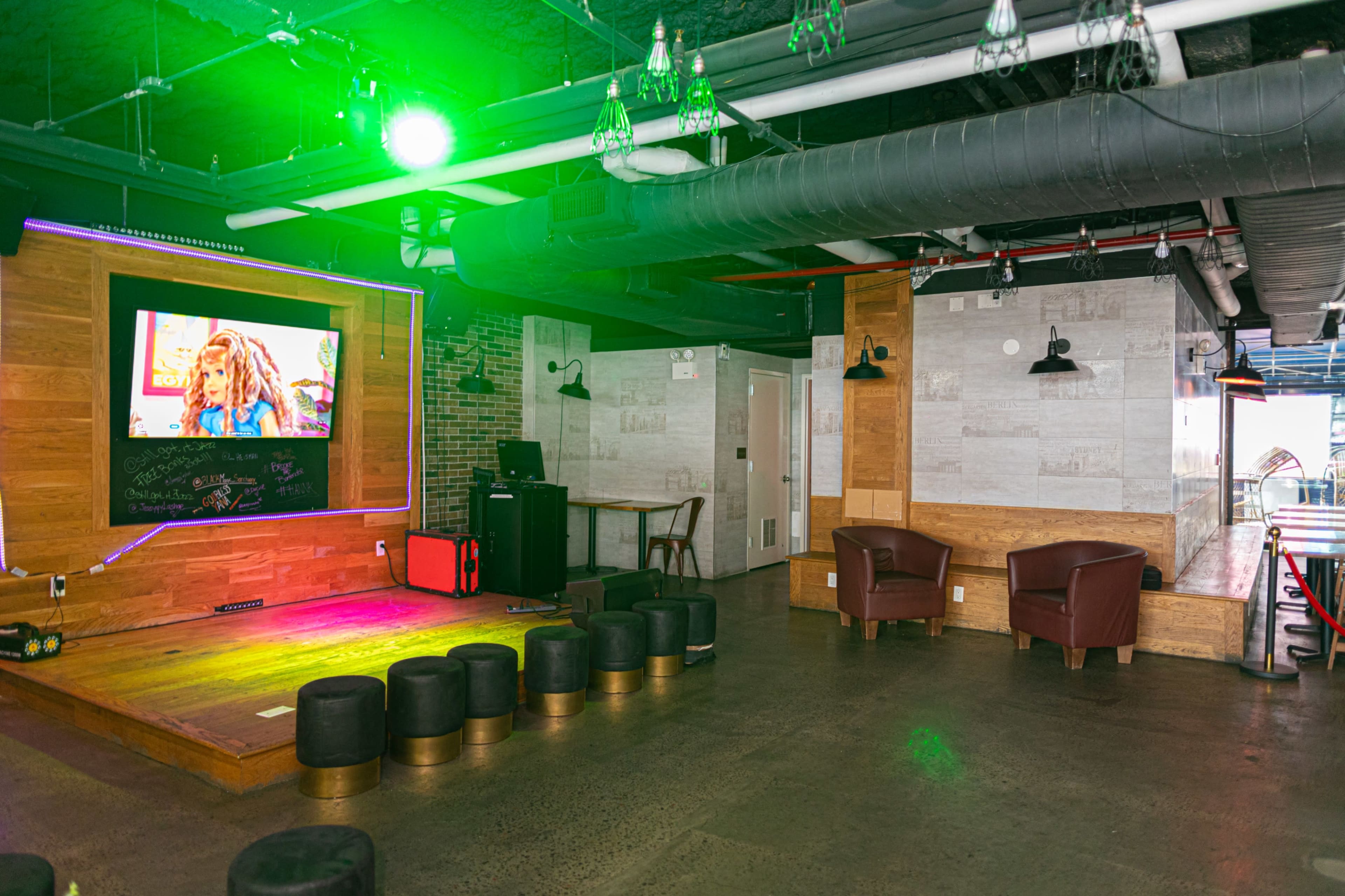 The image shows a modern lounge area with a stage, an entertainment screen displaying a colorful image, and brown leather chairs positioned around the space.