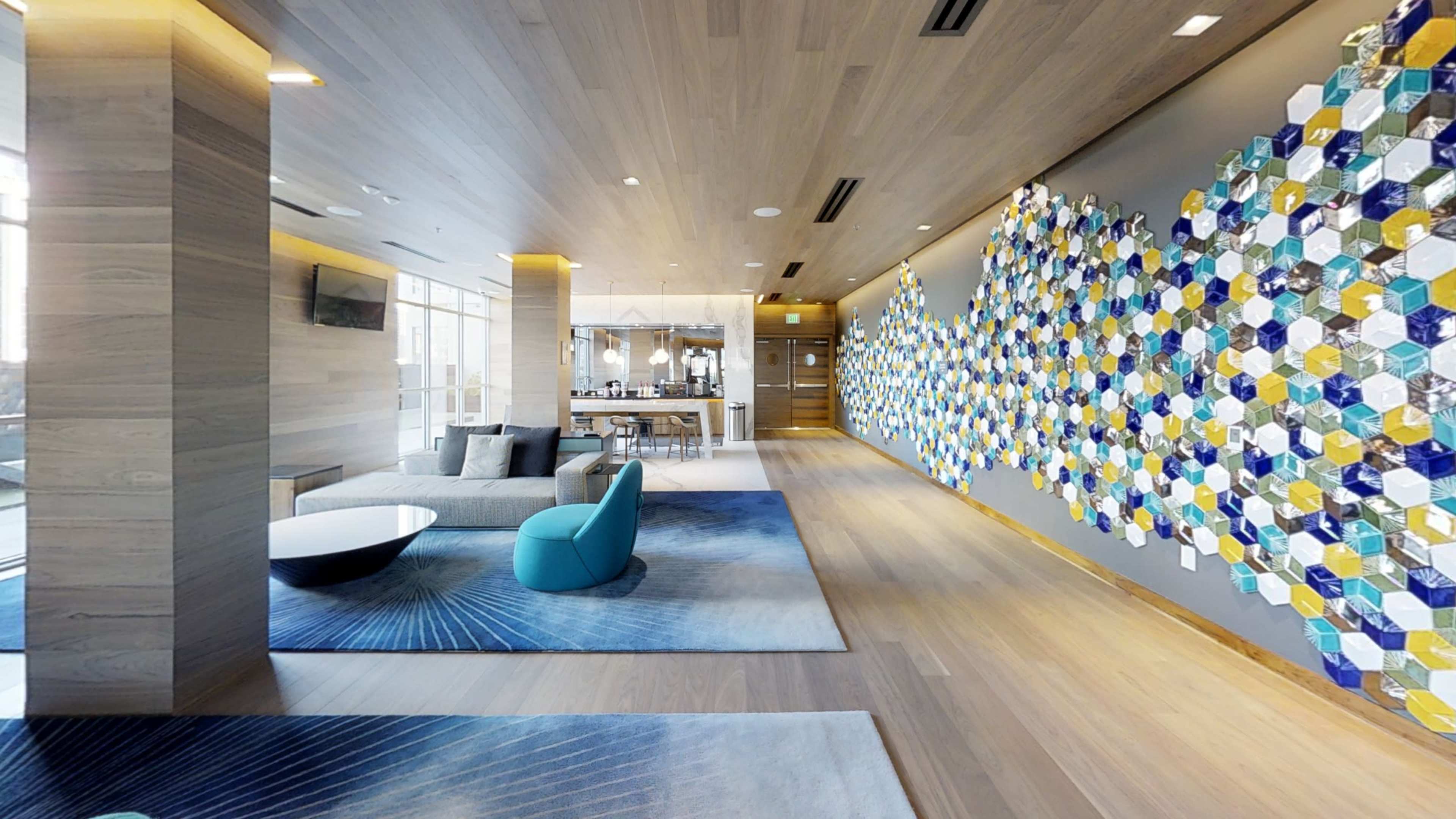 The image shows a modern interior space featuring a long corridor with a blue and yellow hexagonal tile wall, a seating area with a gray sofa and colorful chairs, and an open kitchen visible in the background.