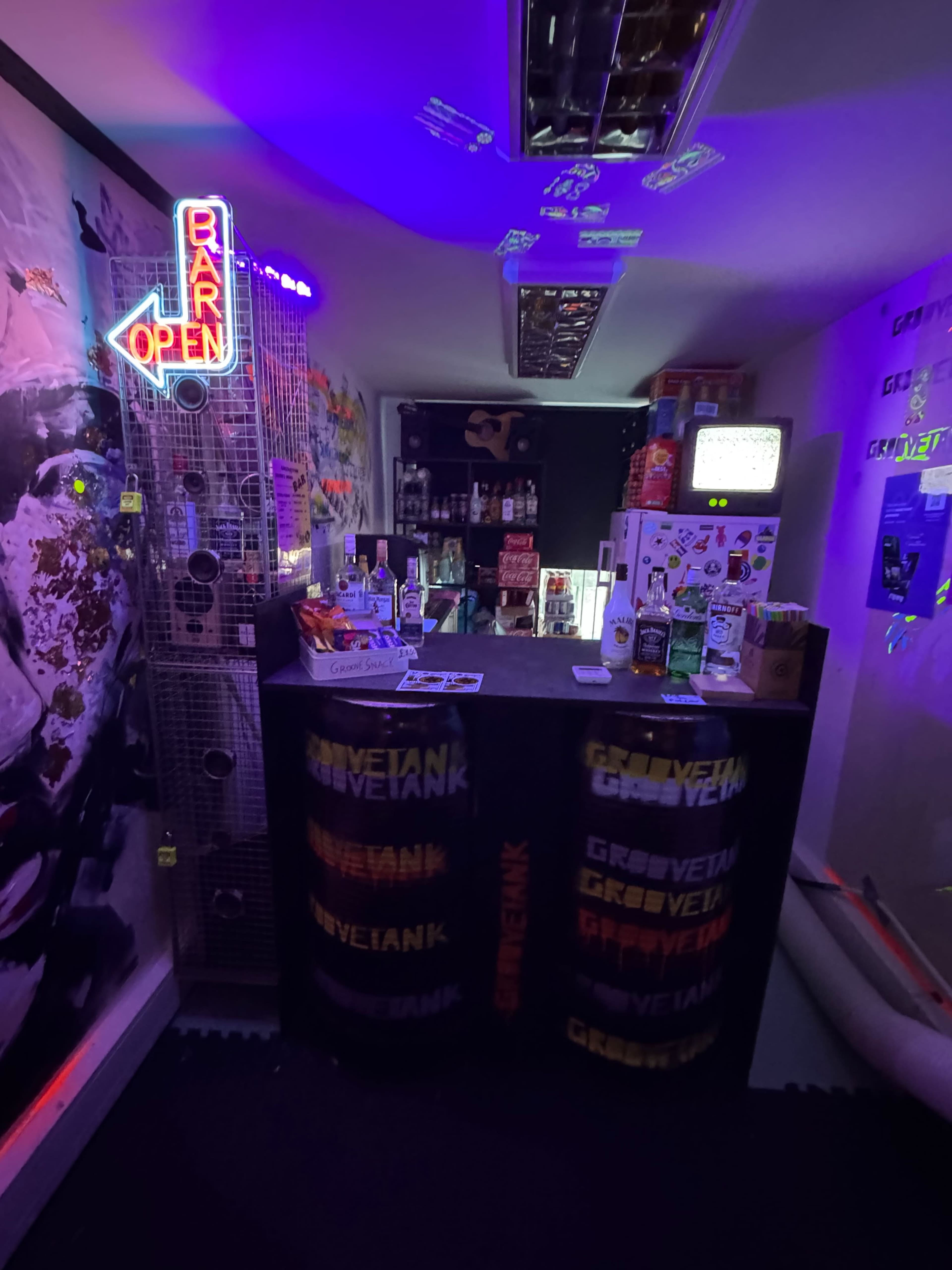 The image shows a small bar area with a neon "BAR OPEN" sign, stocked shelves of drinks, and colorful branding on the counter.