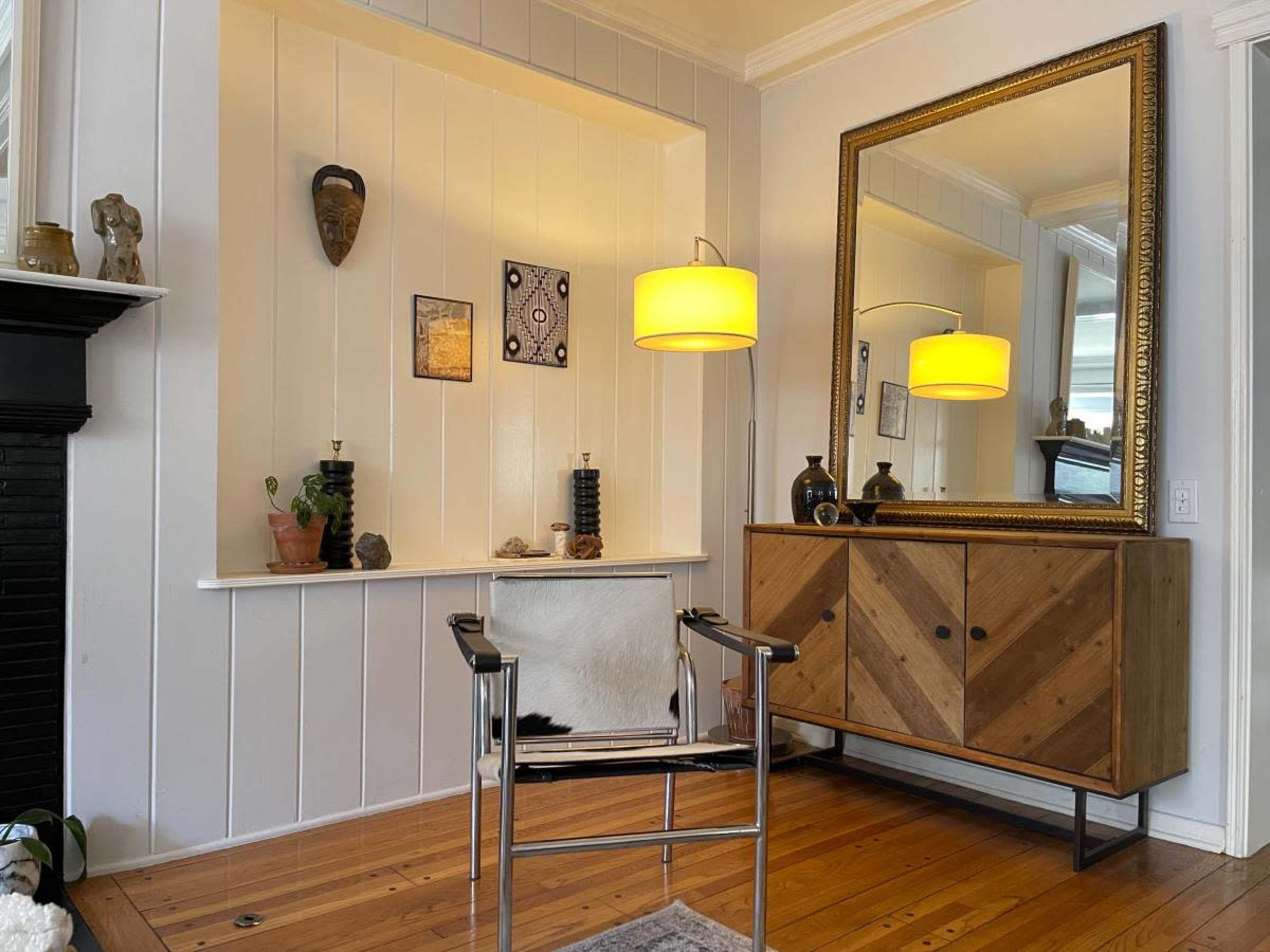 A modern living space features a wooden cabinet, a mirror, a chair, and various decorative items on a shelf against white walls.