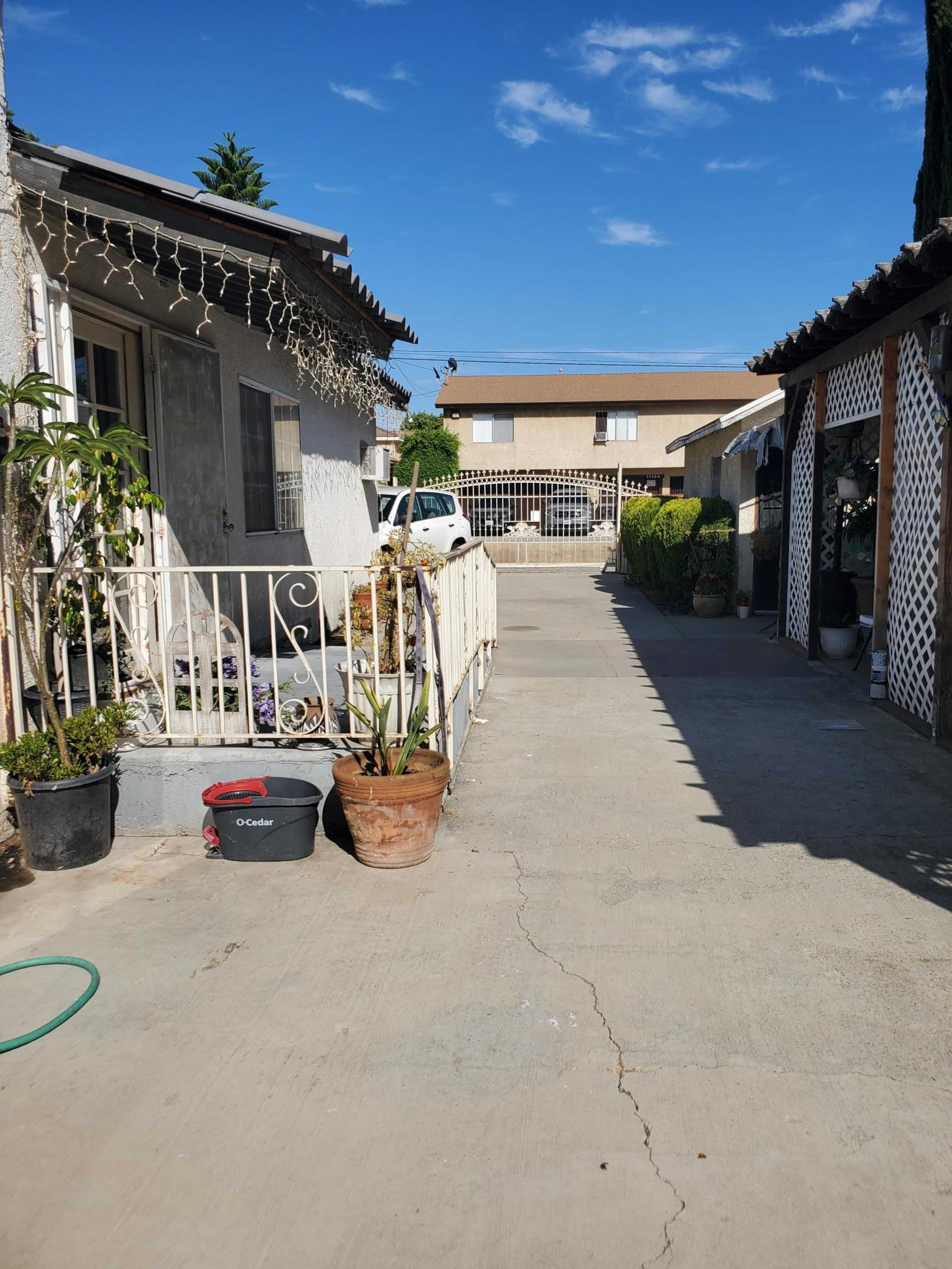 The image shows a concrete pathway leading between two buildings, with plants and a railing on one side and a white vehicle parked in the distance.
