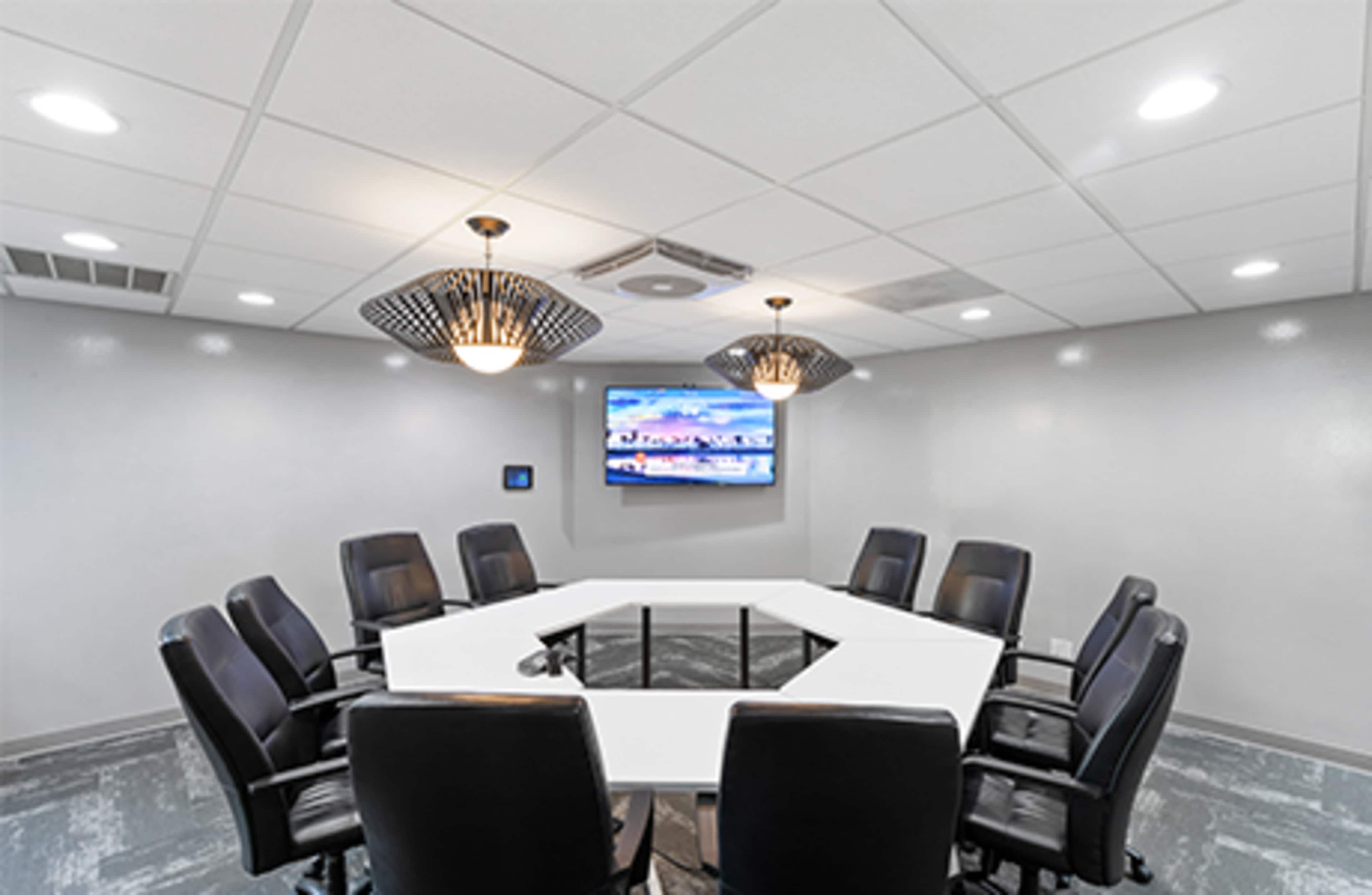 A modern conference room features an octagonal table surrounded by black chairs, with a television mounted on the wall and decorative light fixtures above.