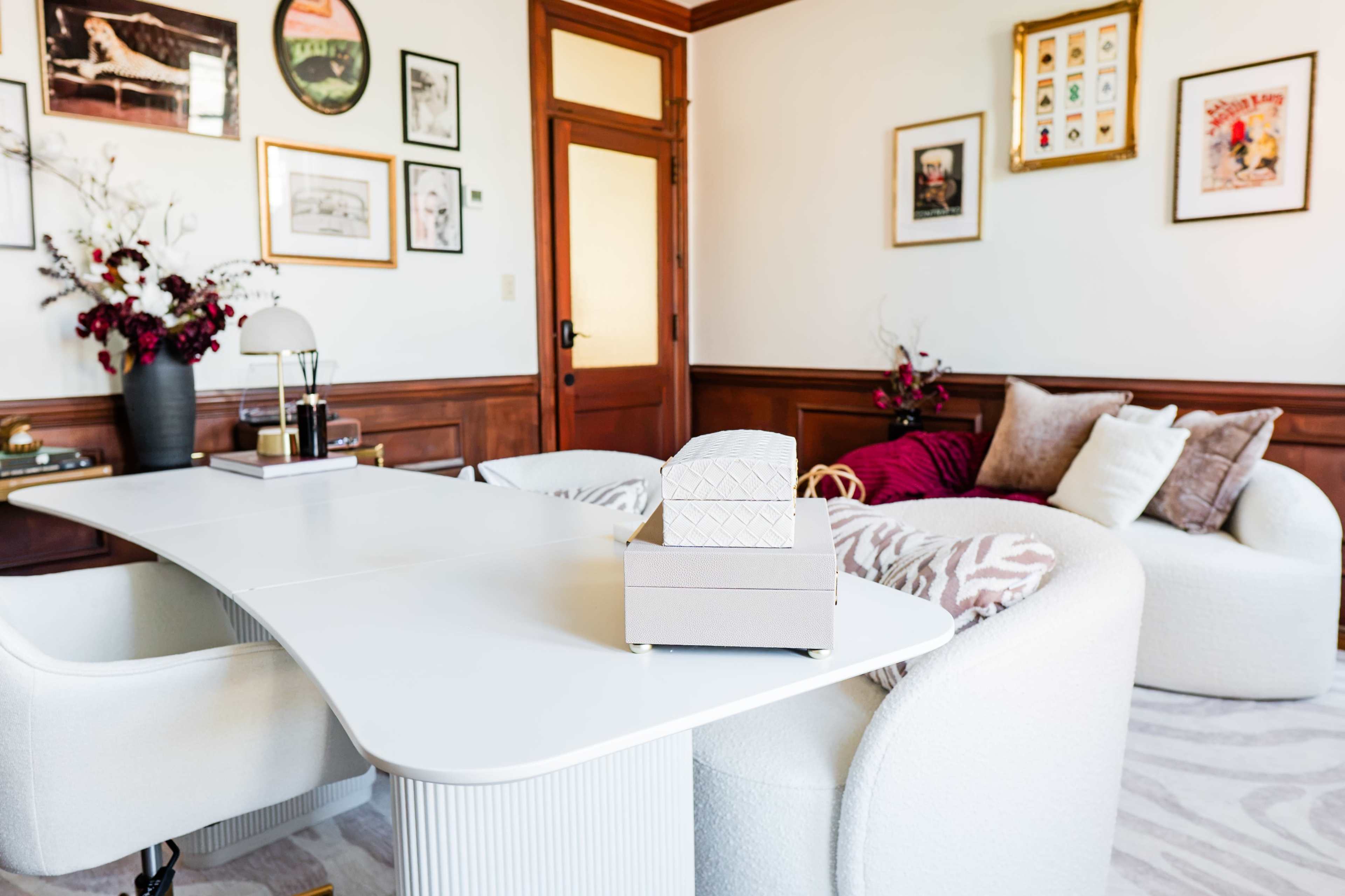 A modern office space features a curved white desk, a cozy sofa with decorative pillows, and framed art on the walls.