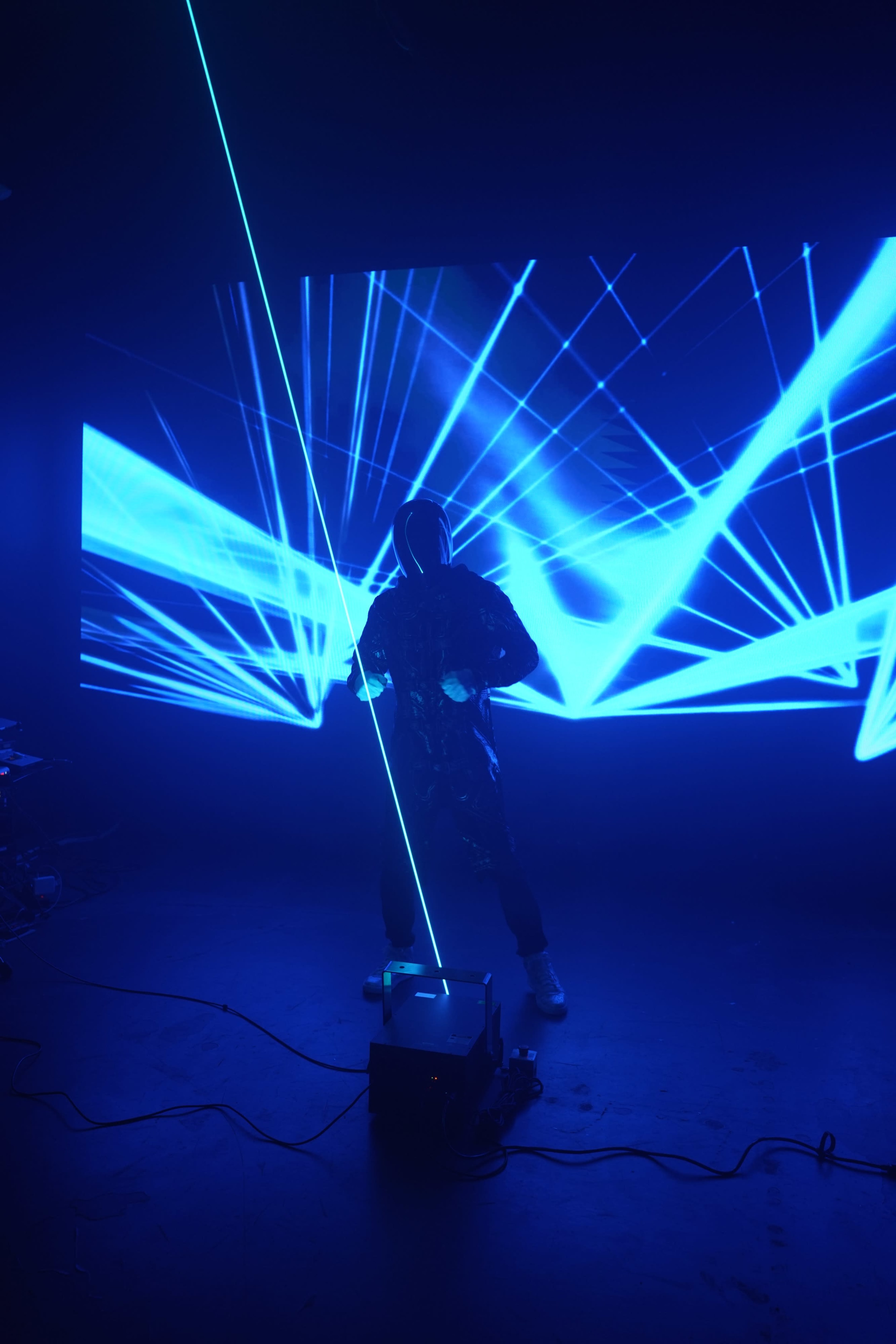 A figure in a dark hooded outfit stands in front of a large screen projecting blue laser beams.