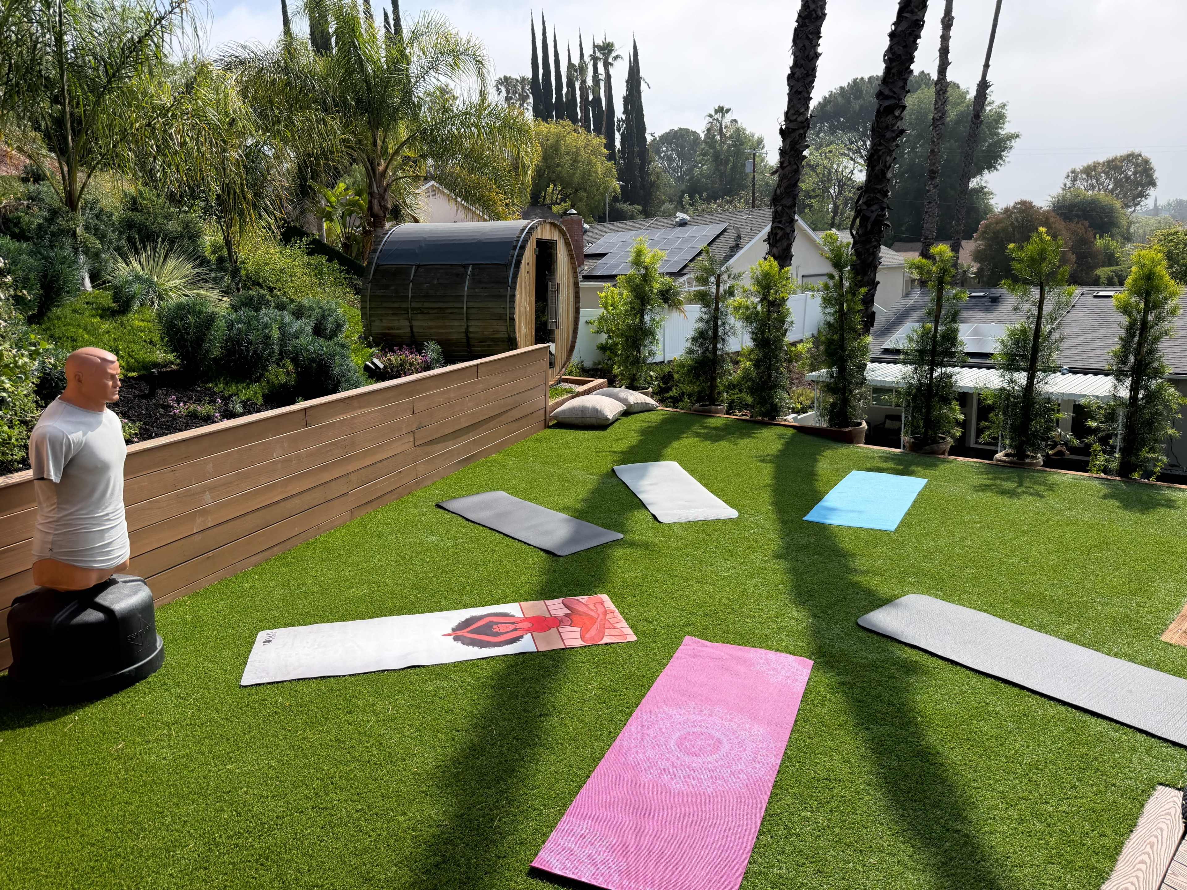 The scene shows a landscaped outdoor area with several yoga mats arranged on artificial grass, surrounded by palm trees and greenery.