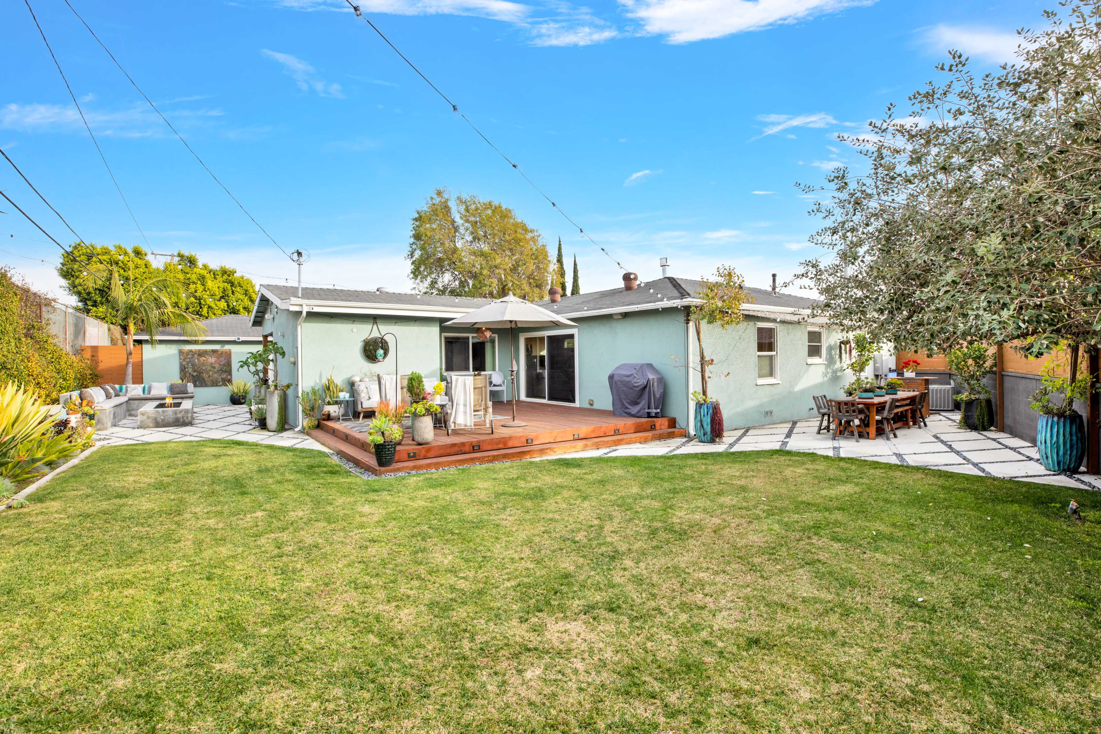 The image shows a backyard featuring a patio area with a table, an umbrella, and landscaped grass surrounded by several trees and decorative plants.