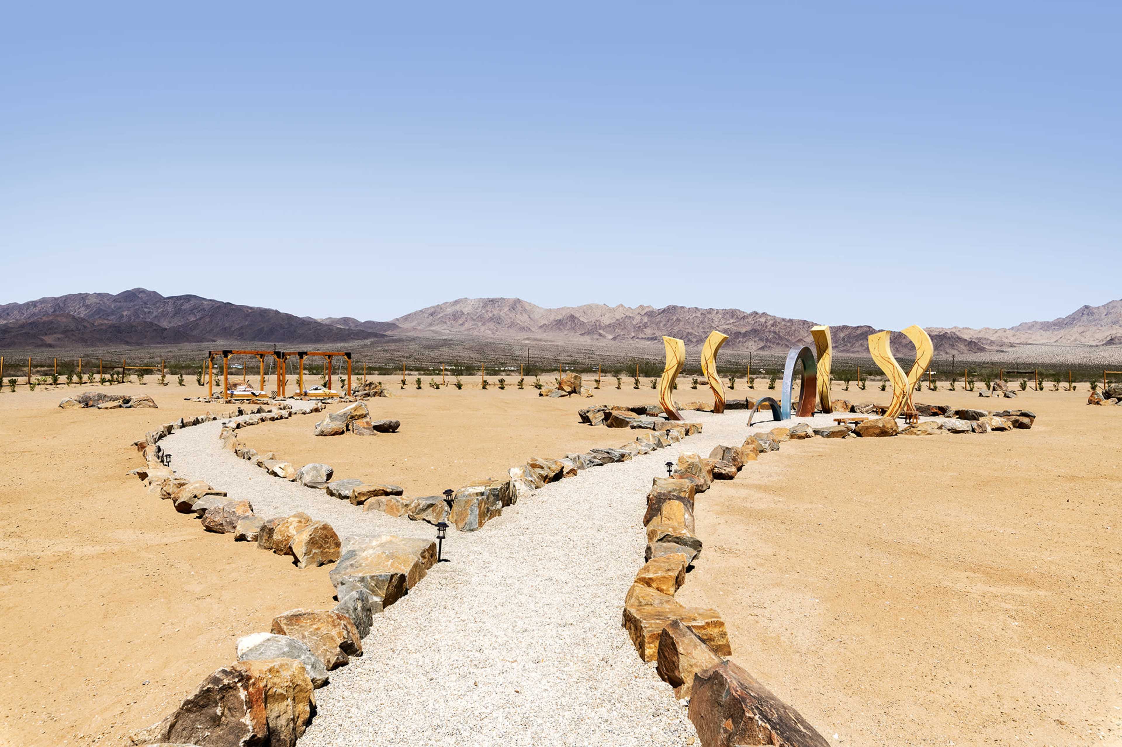 A gravel pathway winds through a desert landscape, leading to a series of sculptural structures and mountains in the background.