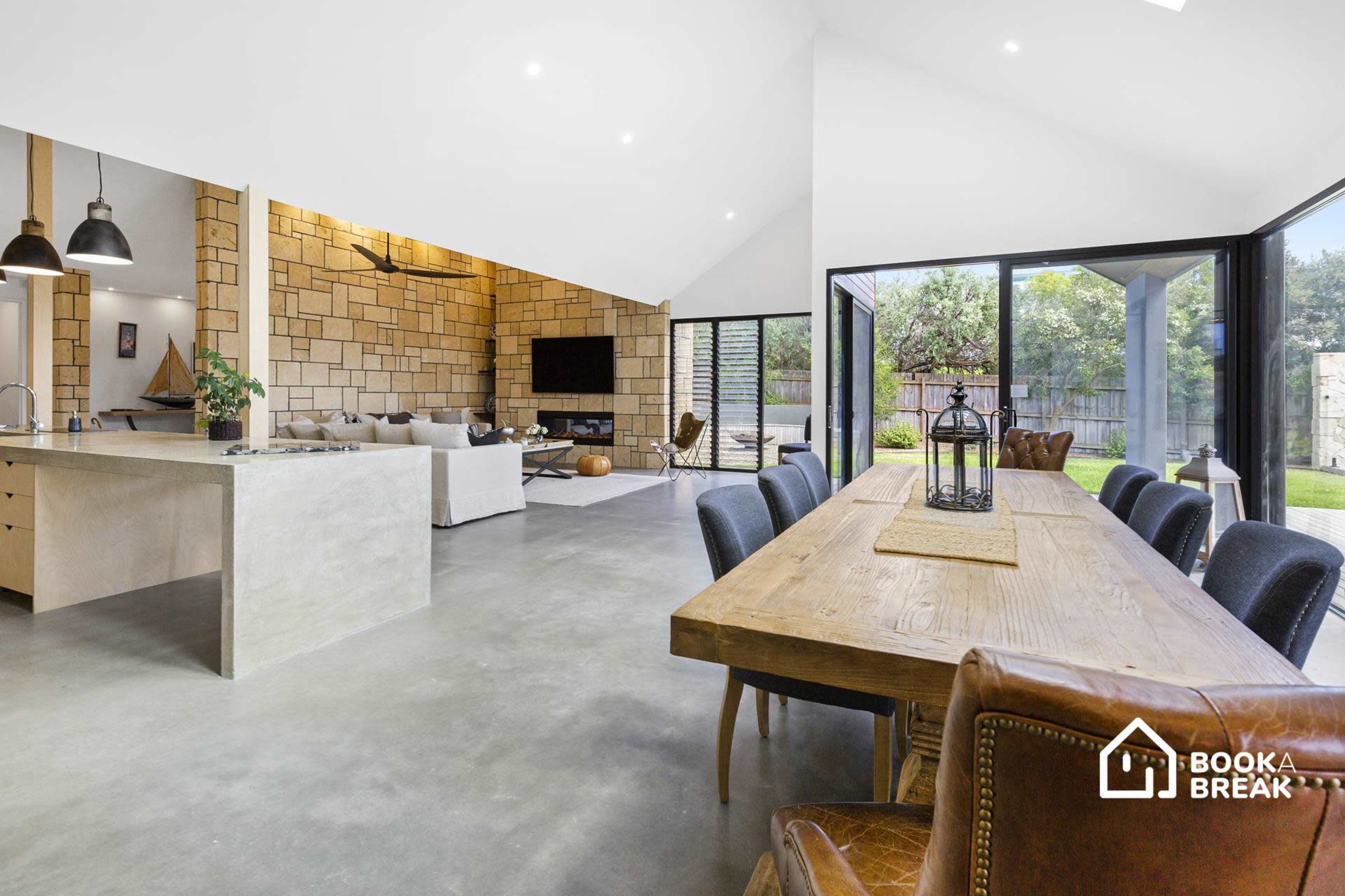 A spacious, modern open-concept living and dining area with concrete flooring, large windows, and a wooden dining table.