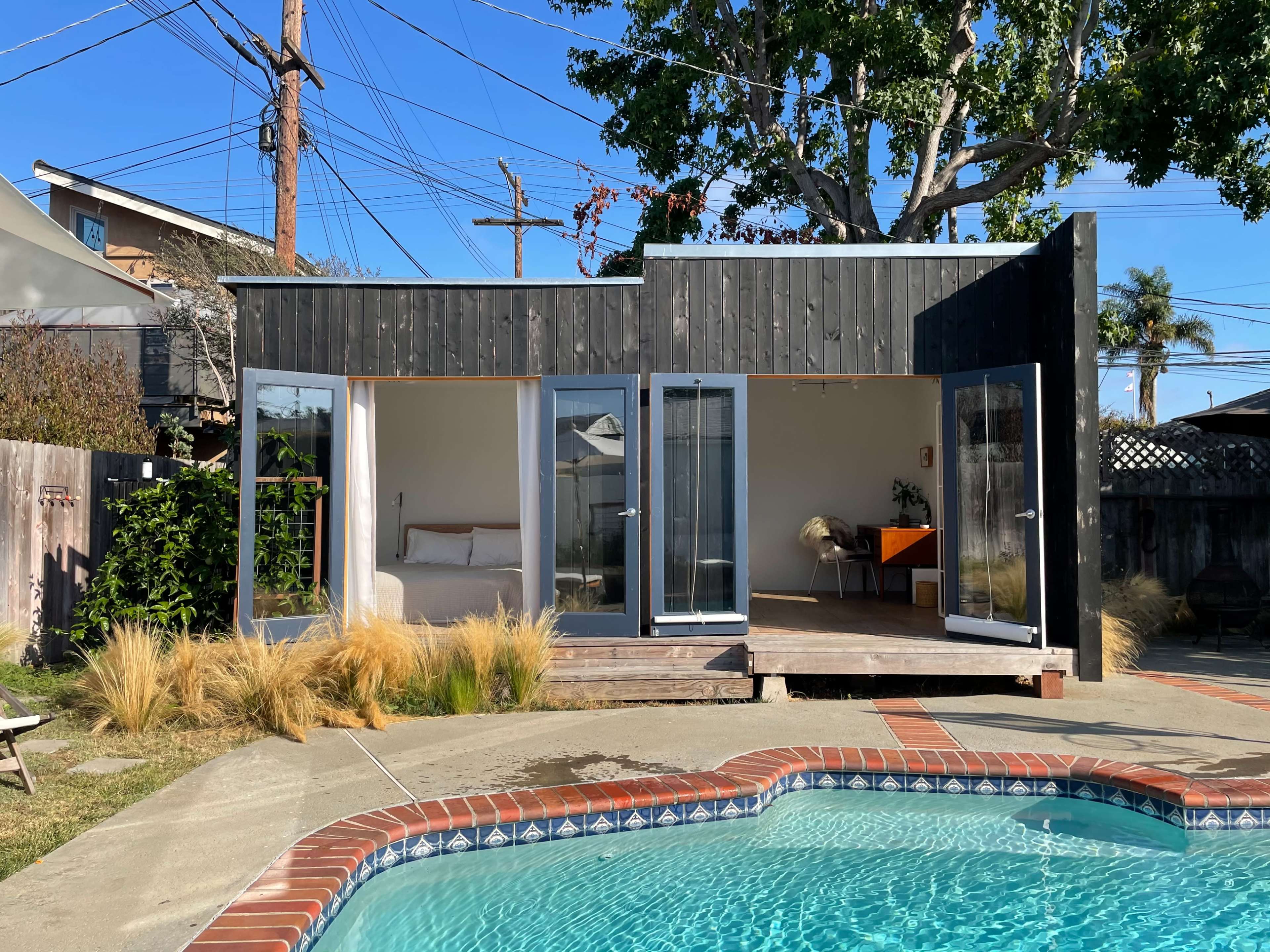 A small modern structure with large glass doors sits beside a swimming pool, surrounded by greenery and utility poles against a clear blue sky.