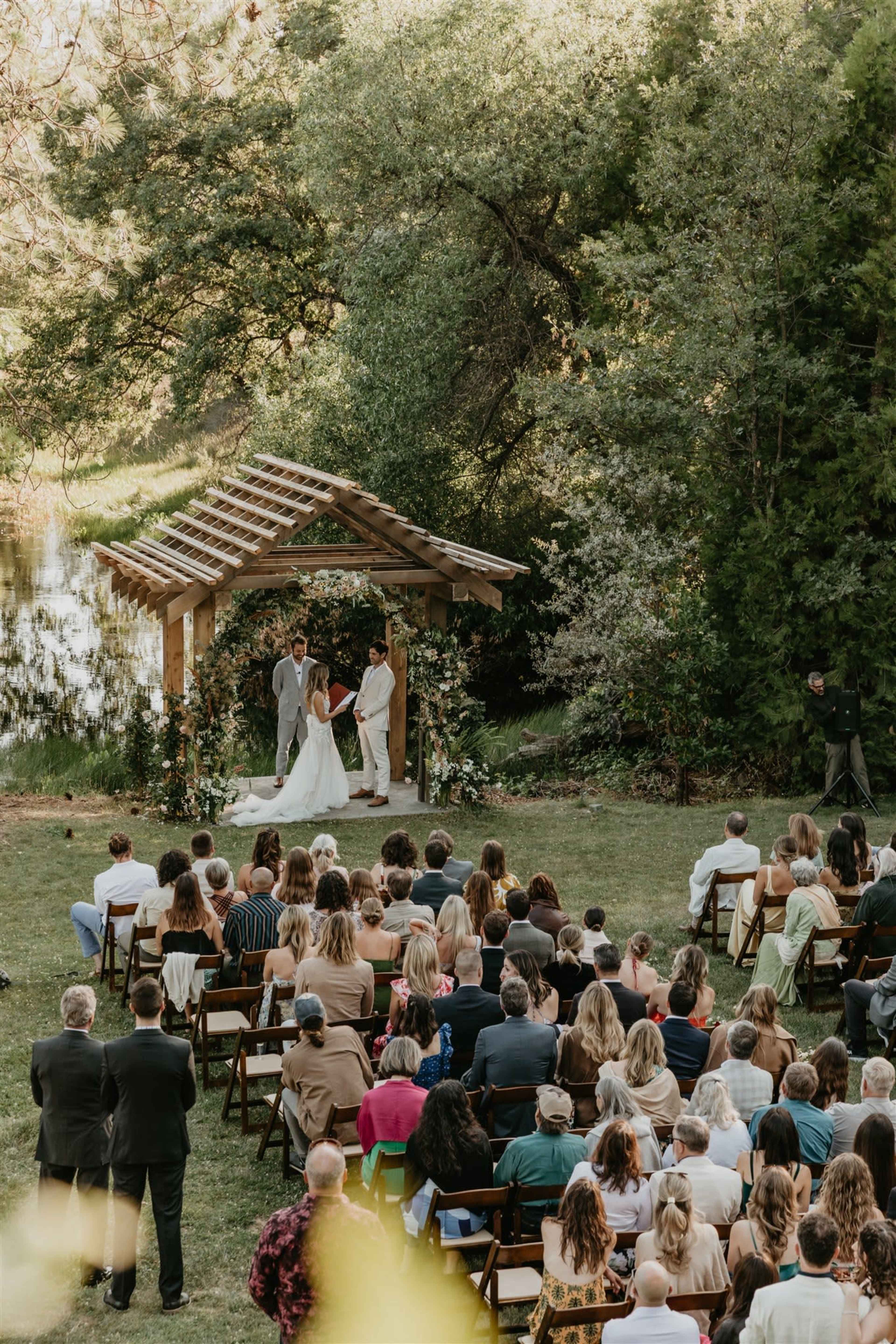 A couple is exchanging vows under a wooden arbor adorned with greenery while attendees sit in rows of chairs nearby.