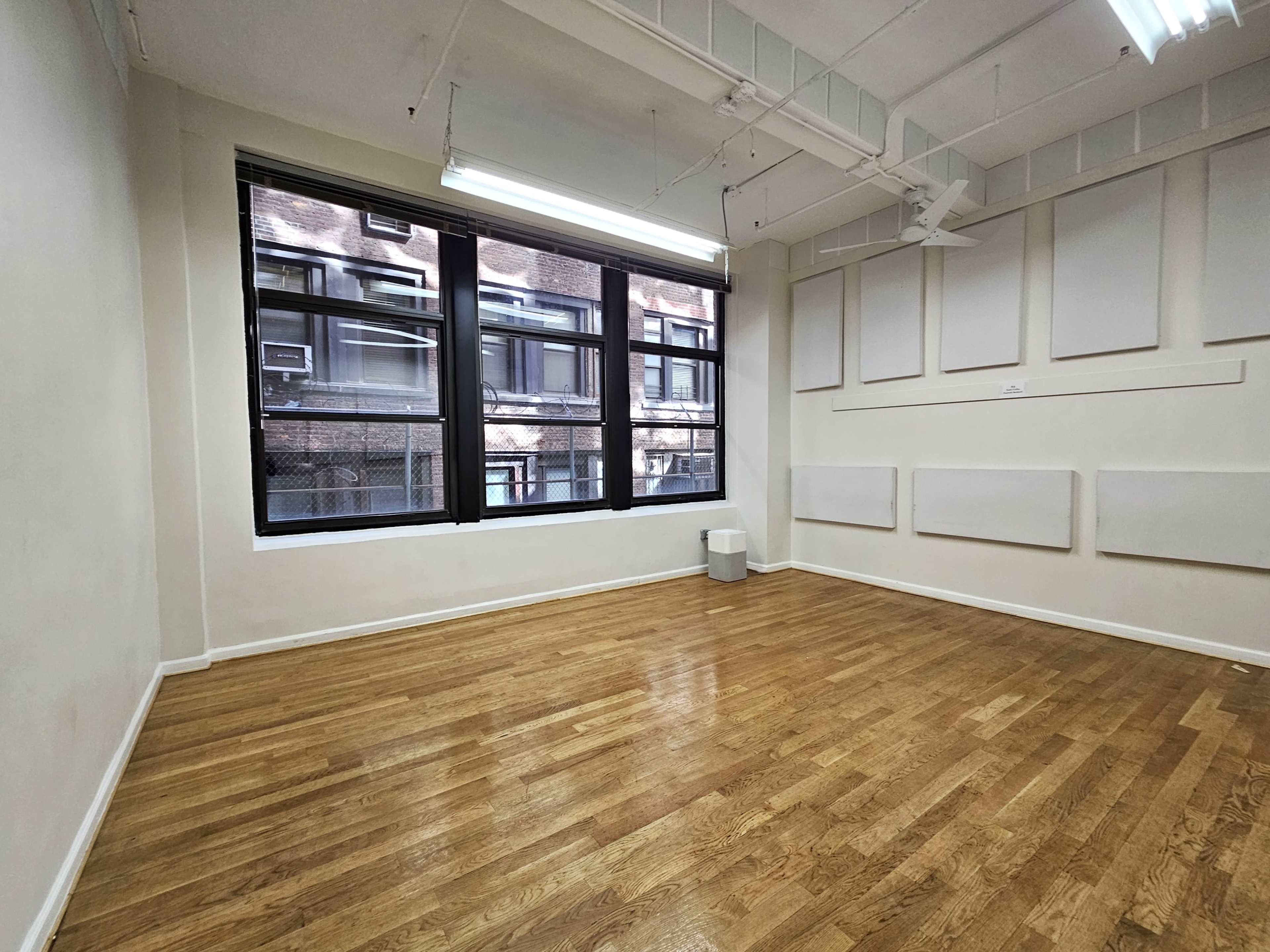 The image shows a vacant room with wooden flooring, large windows, and bare walls.