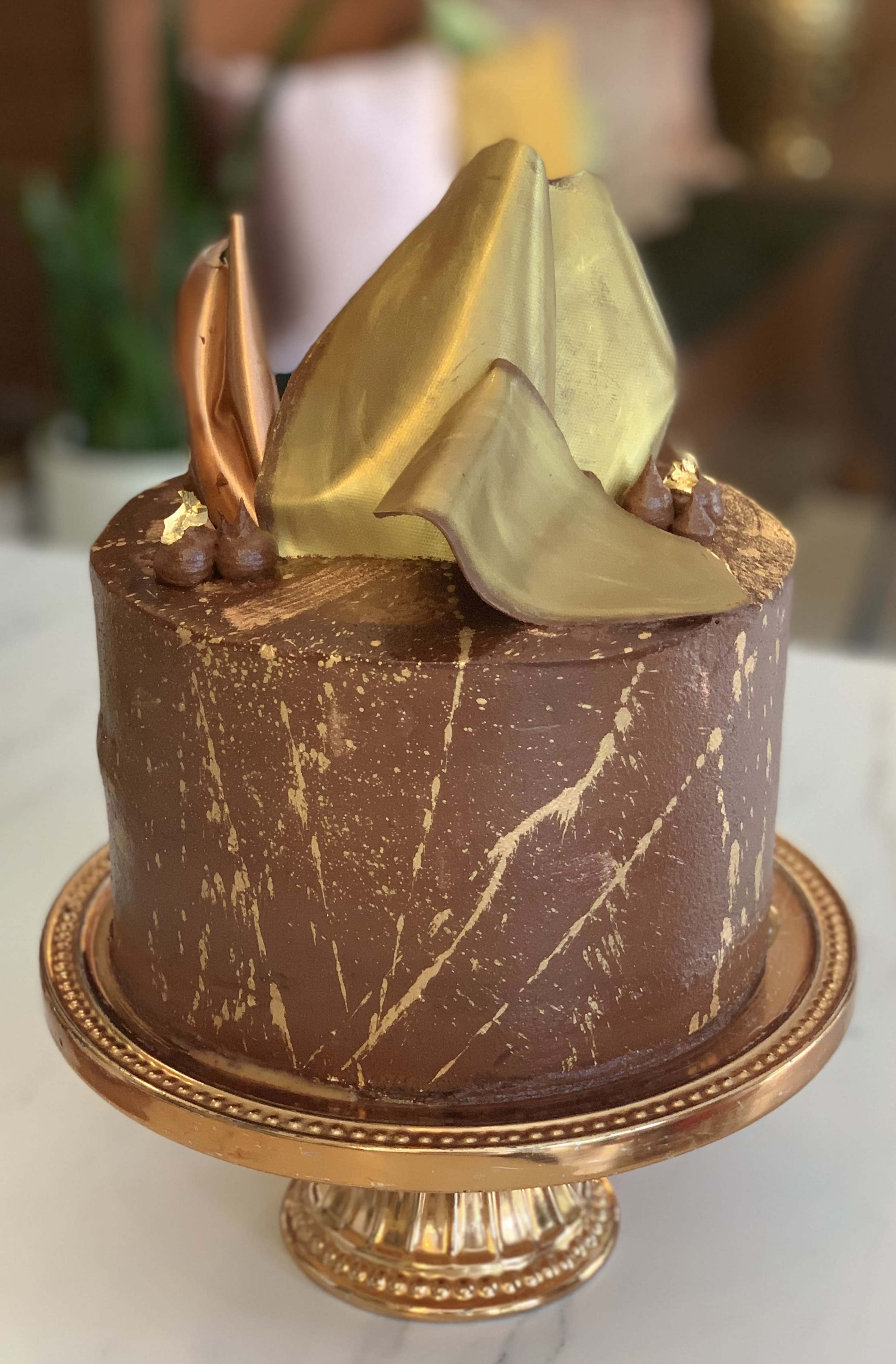 The image shows a chocolate cake decorated with gold and copper accents on a decorative golden stand.