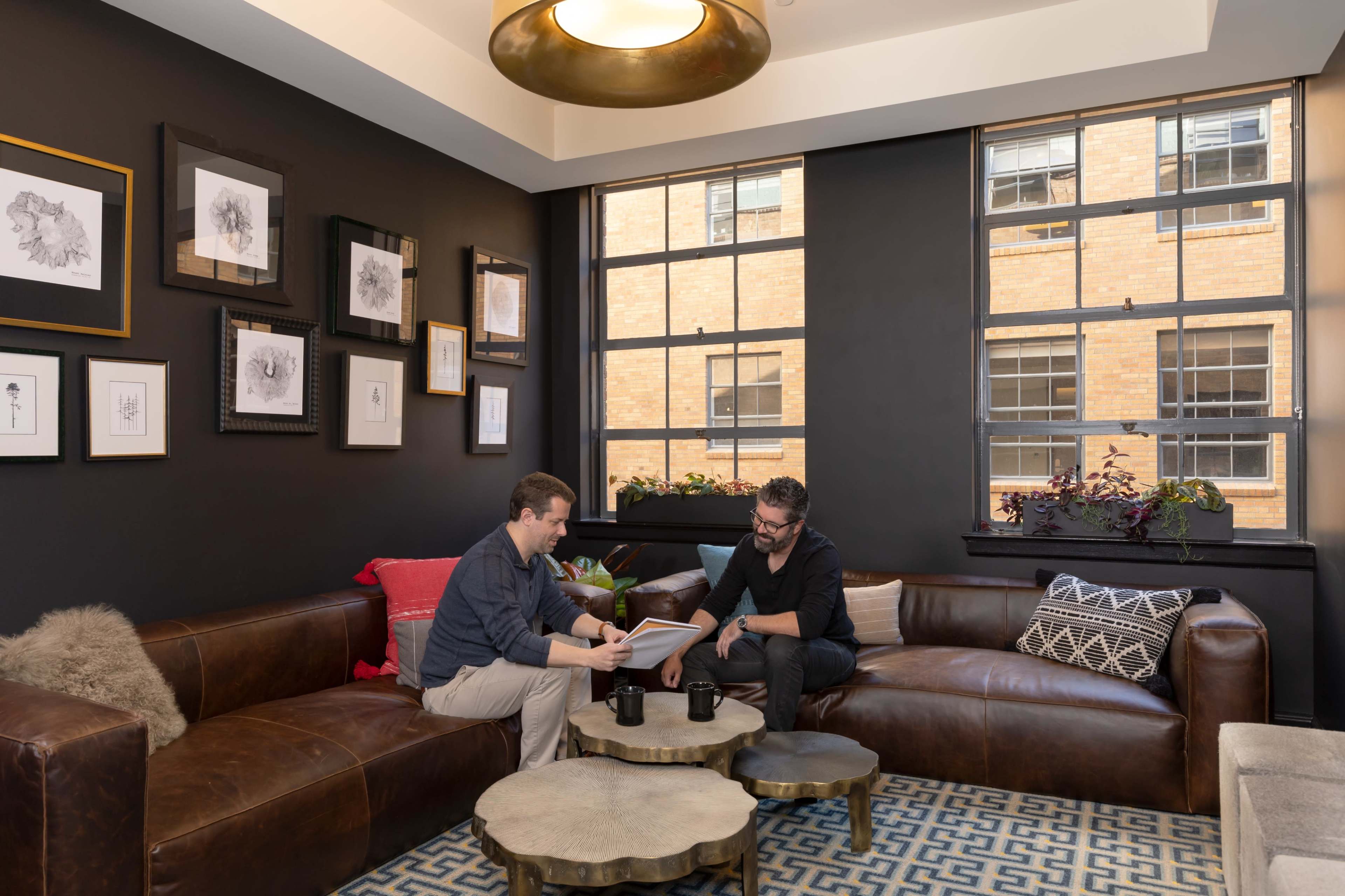 Two men sit on a brown leather couch in a well-lit room with large windows, discussing a document while surrounded by framed artwork on the walls.