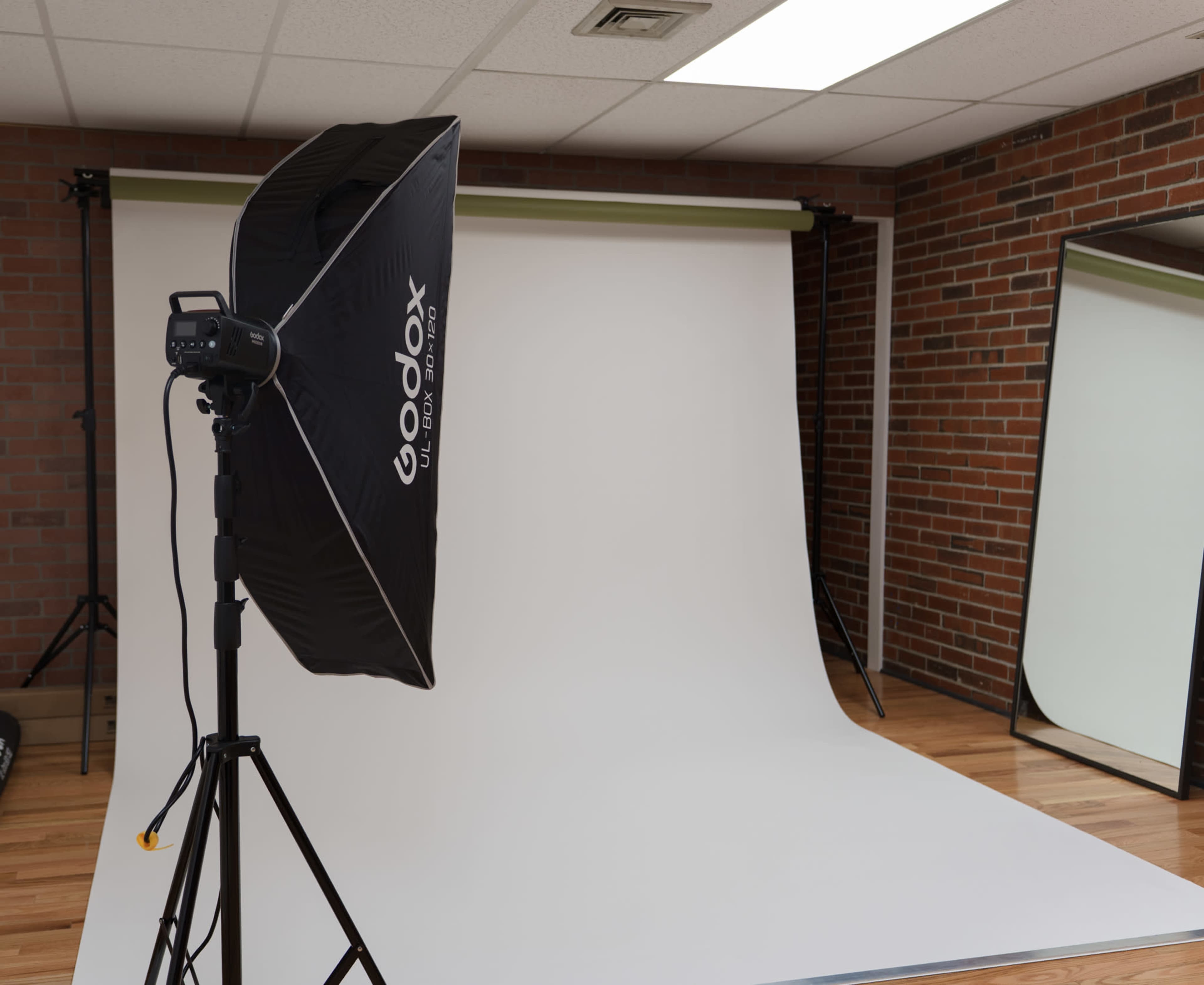 The image shows a photography studio setup with a softbox light, a white backdrop, and a mirror against a brick wall.