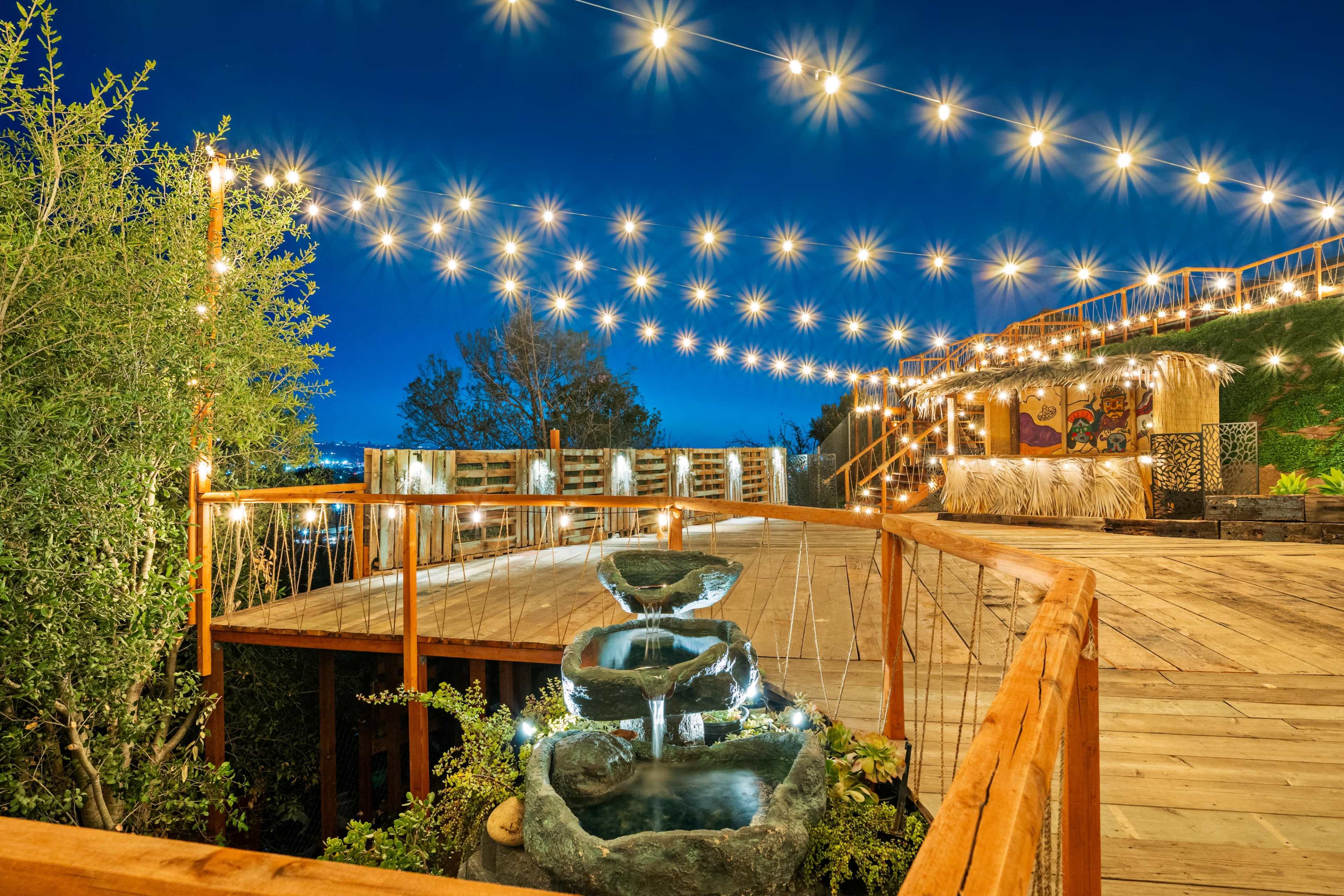 A wooden deck features a tiered water fountain illuminated by lights, surrounded by lush greenery and strung lights overhead in the evening sky.