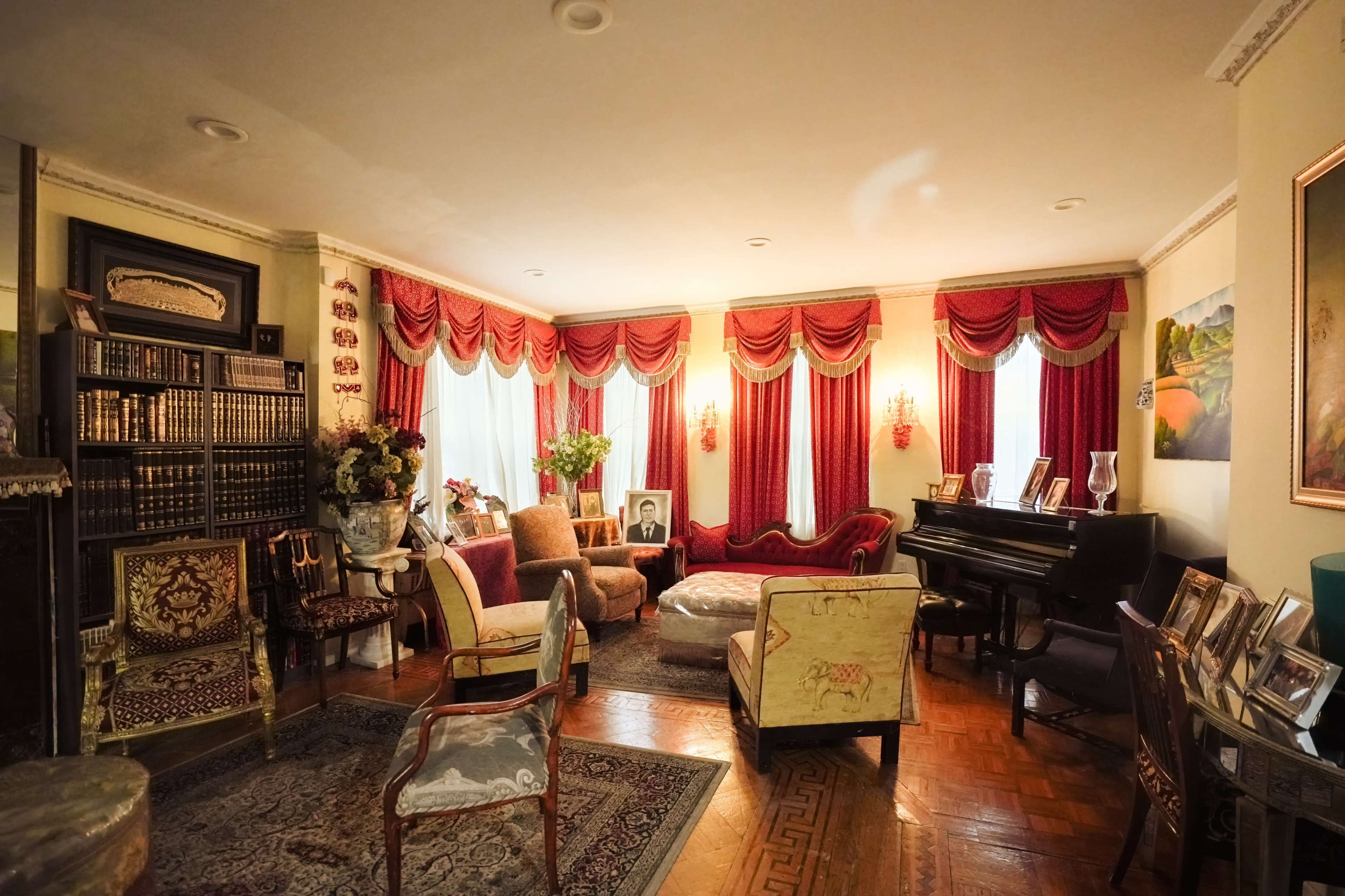 The image shows a cozy living room with plush seating, a grand piano, and a bookshelf filled with books, all adorned with elegant curtains and decor.