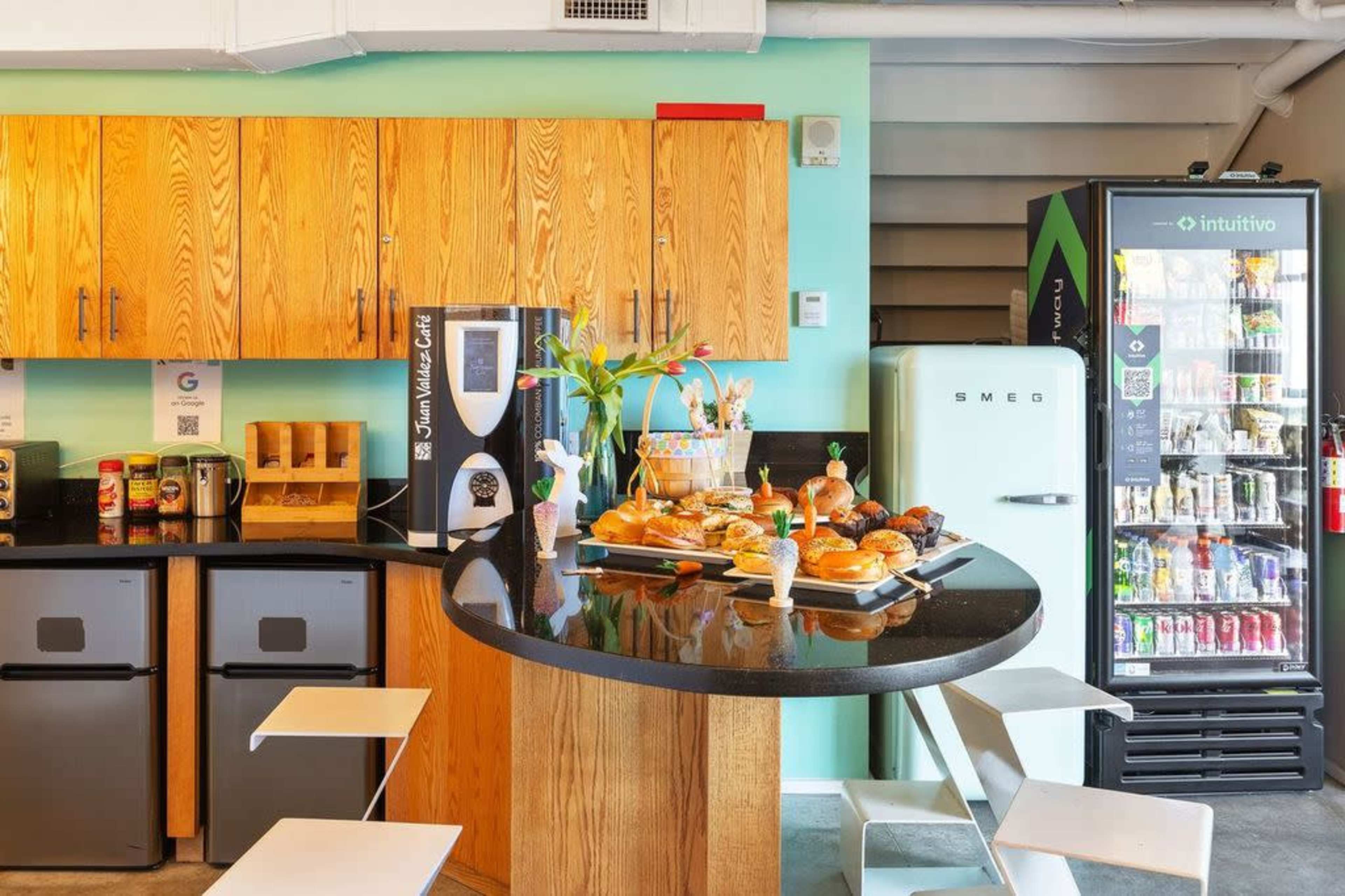 A modern kitchen space features wooden cabinets, a coffee machine, a display of baked goods on a countertop, and a sleek green refrigerator beside a vending machine.
