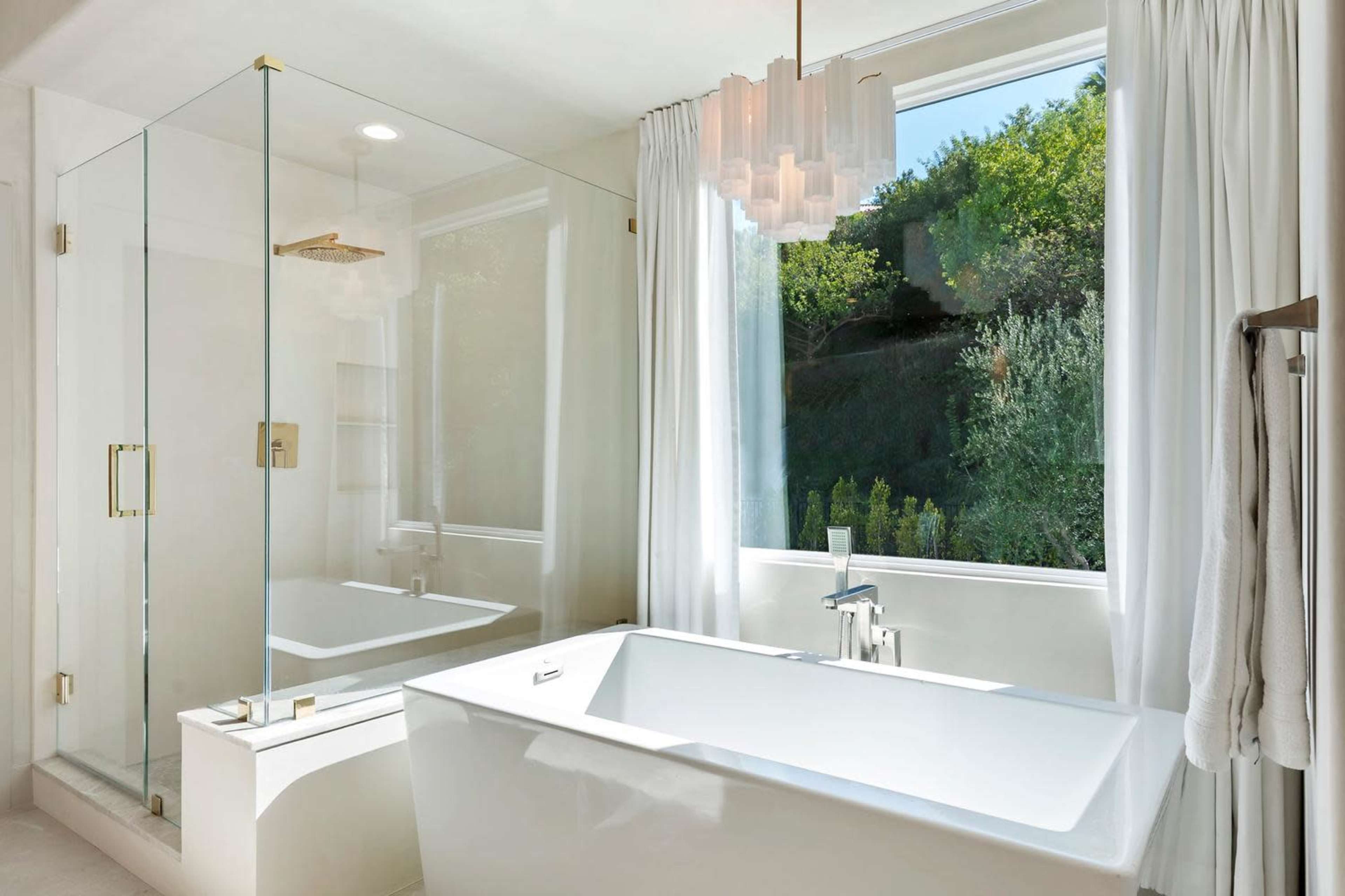 The image shows a modern bathroom featuring a glass shower enclosure, a freestanding bathtub, and a large window with natural light illuminating the space.