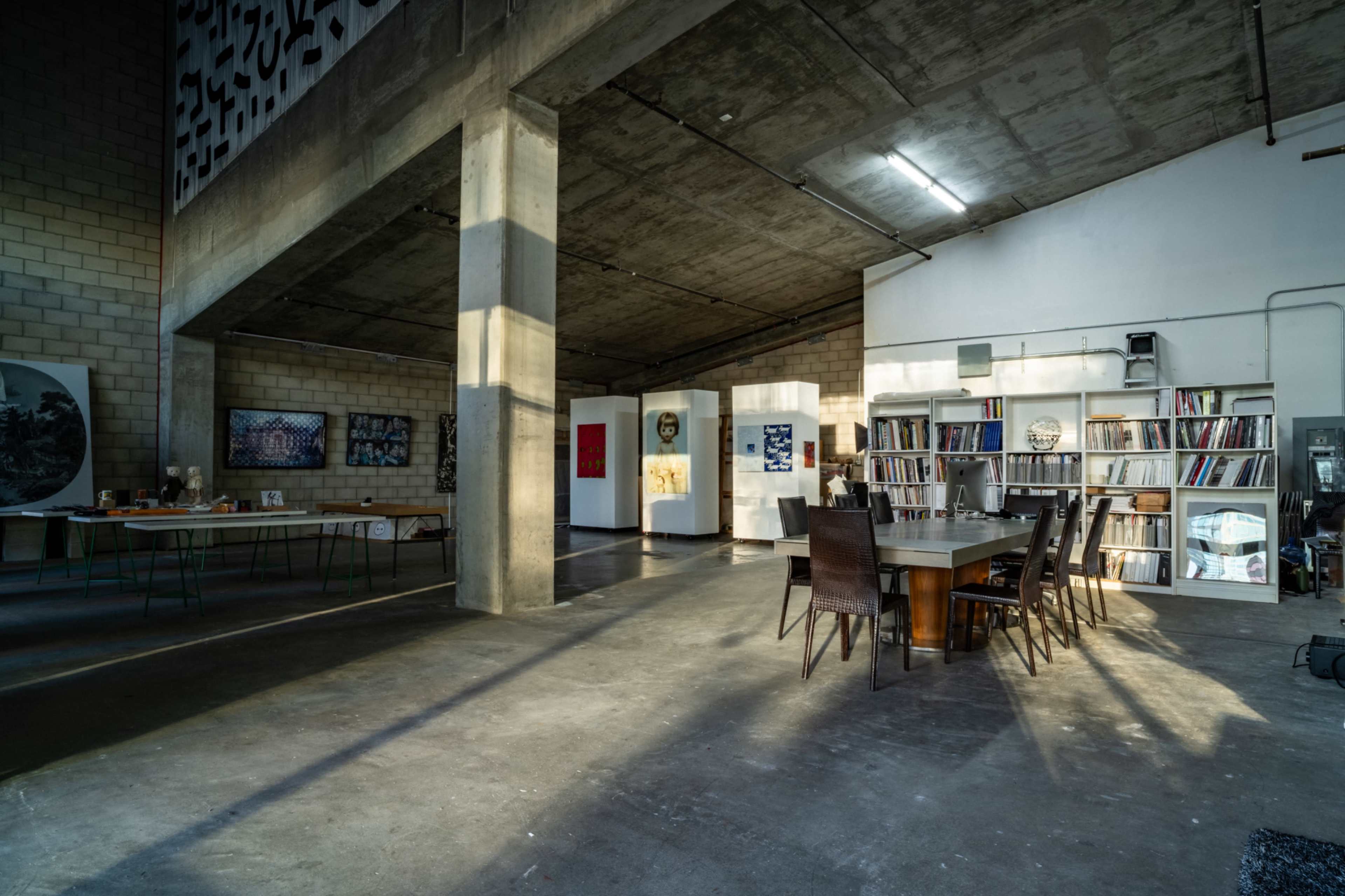 The image shows a spacious industrial-style room with a large wooden dining table surrounded by chairs, set against a backdrop of concrete walls and shelves filled with books and art.