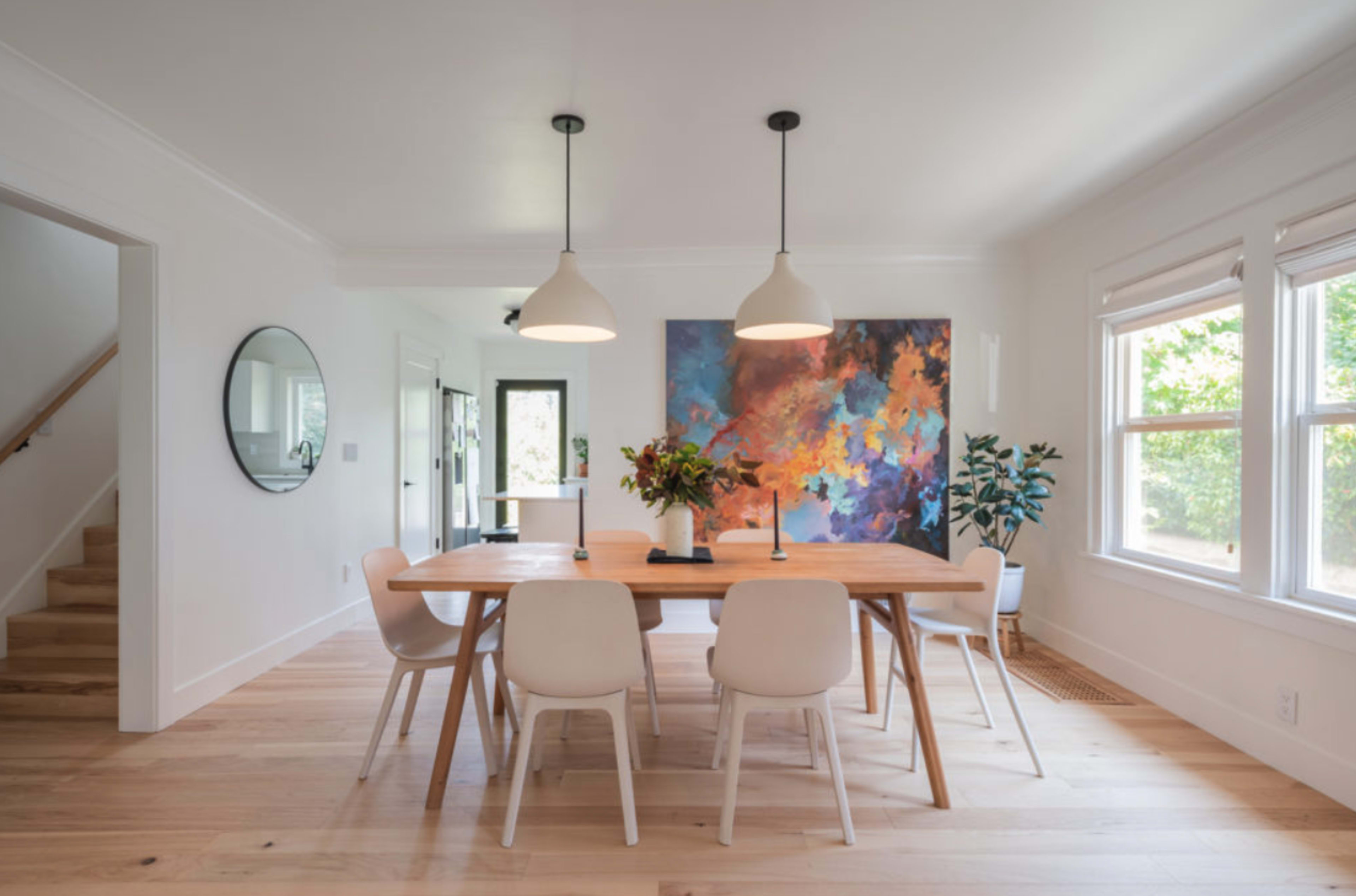 A spacious dining room features a wooden table surrounded by white chairs, with a large colorful artwork on the wall and natural light illuminating the space.