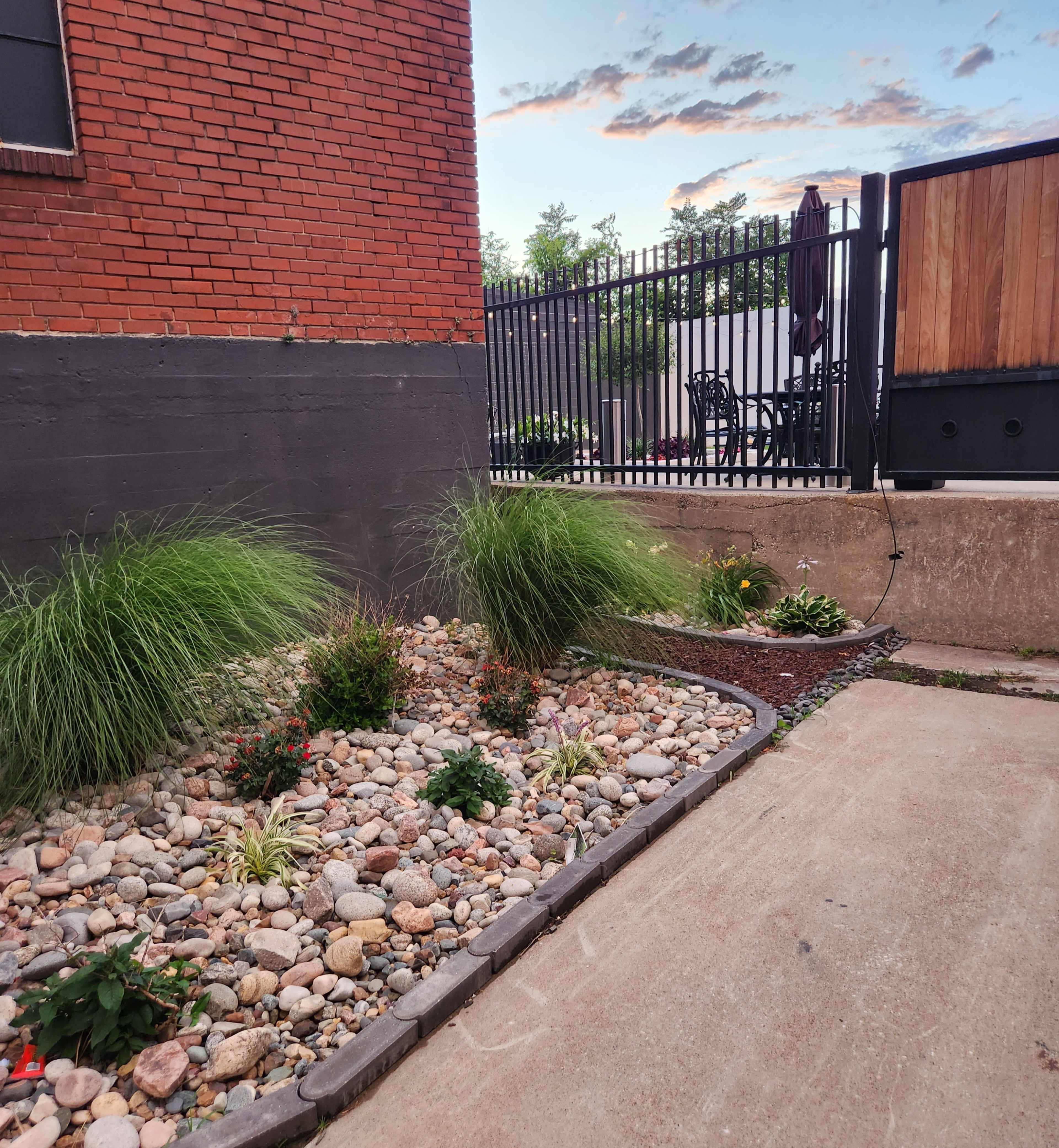 The scene features a landscaped area with a gravel bed, bordered by stones, and adorned with shrubs and flowers next to a paved path and a fenced seating area in the background.