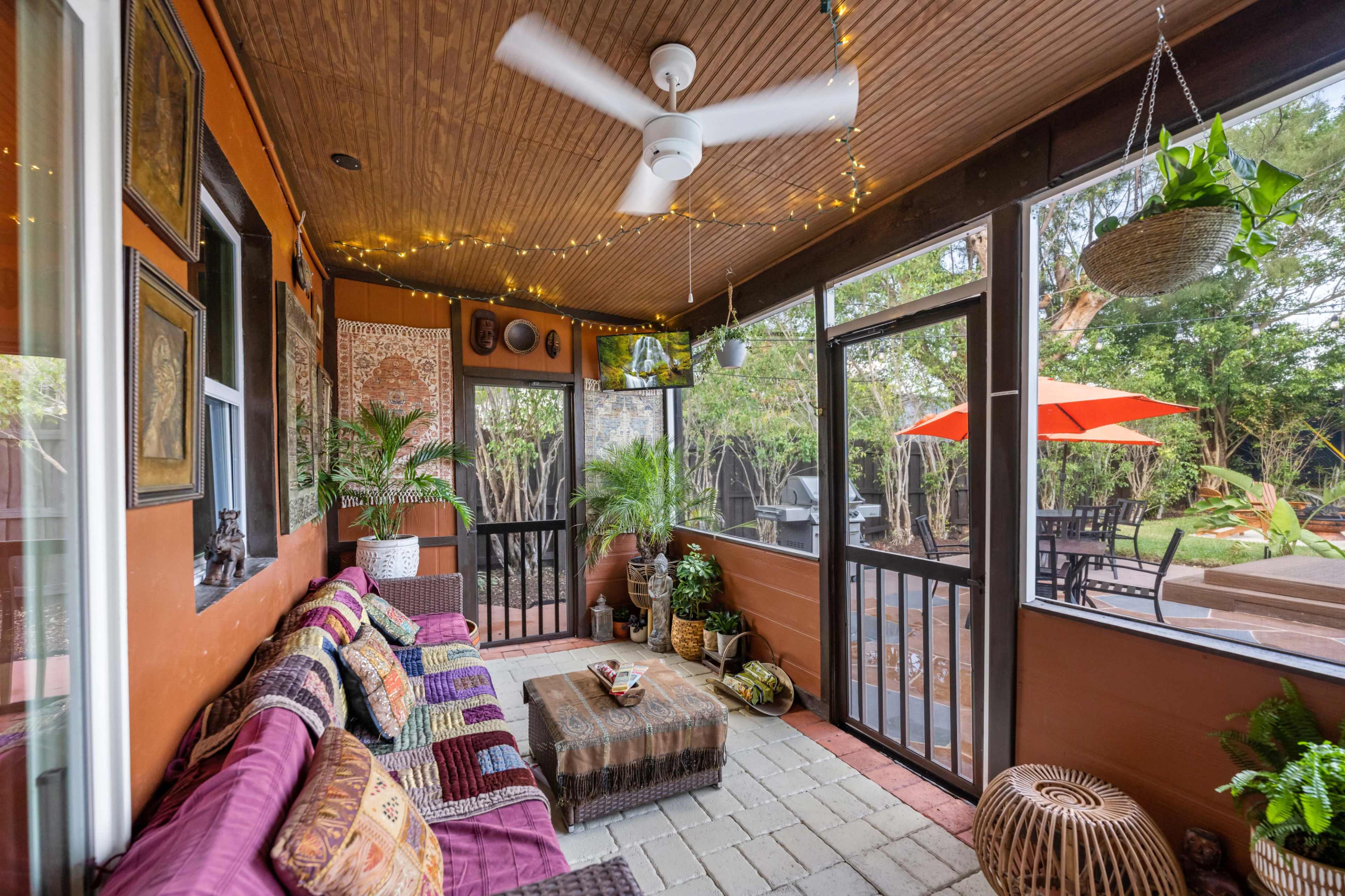 A screened-in porch featuring a seating area with colorful cushions, a coffee table, and potted plants, illuminated by string lights.