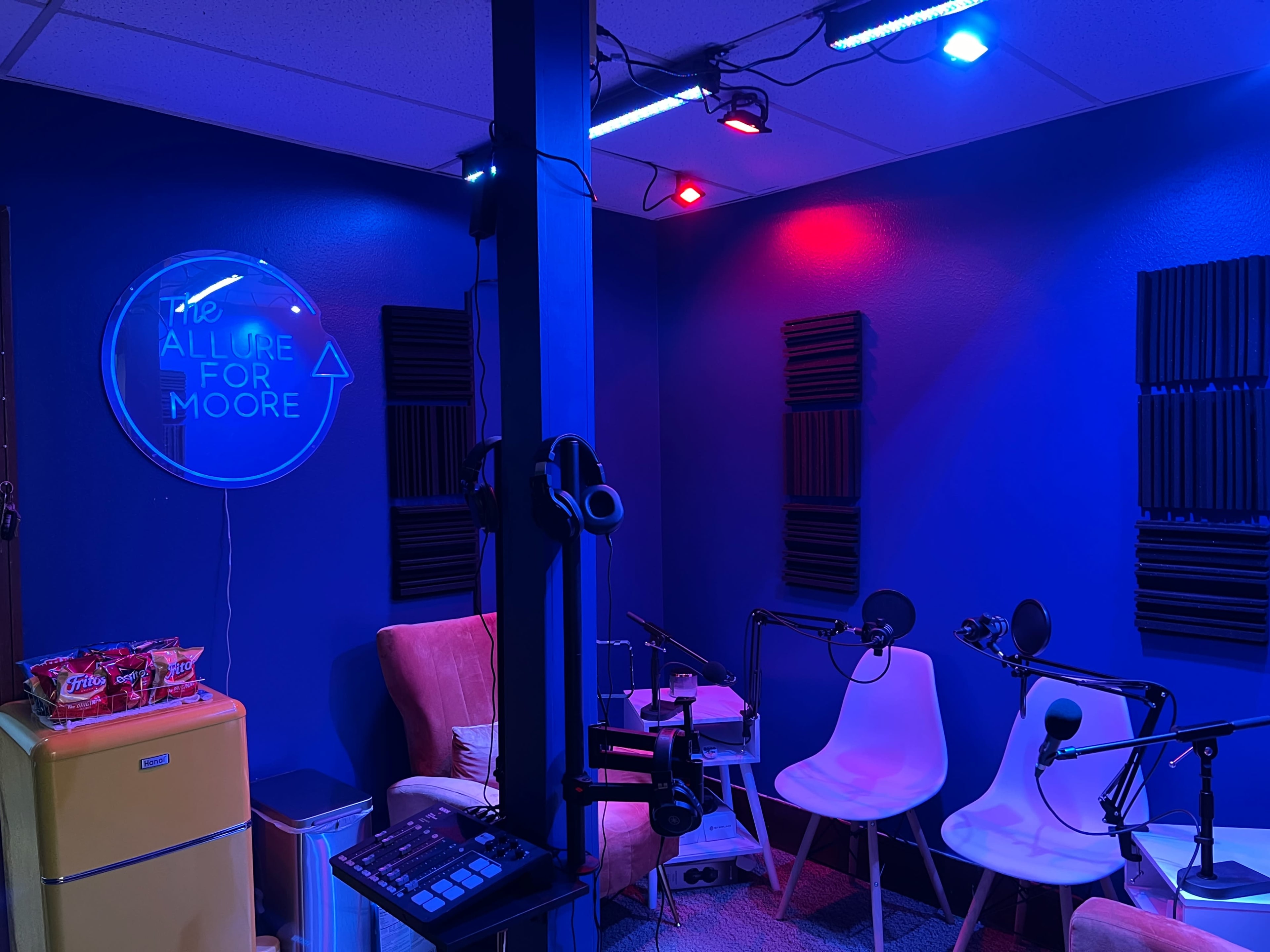 A recording studio with two microphones, white chairs, soundproofing panels on the walls, and colorful lighting.