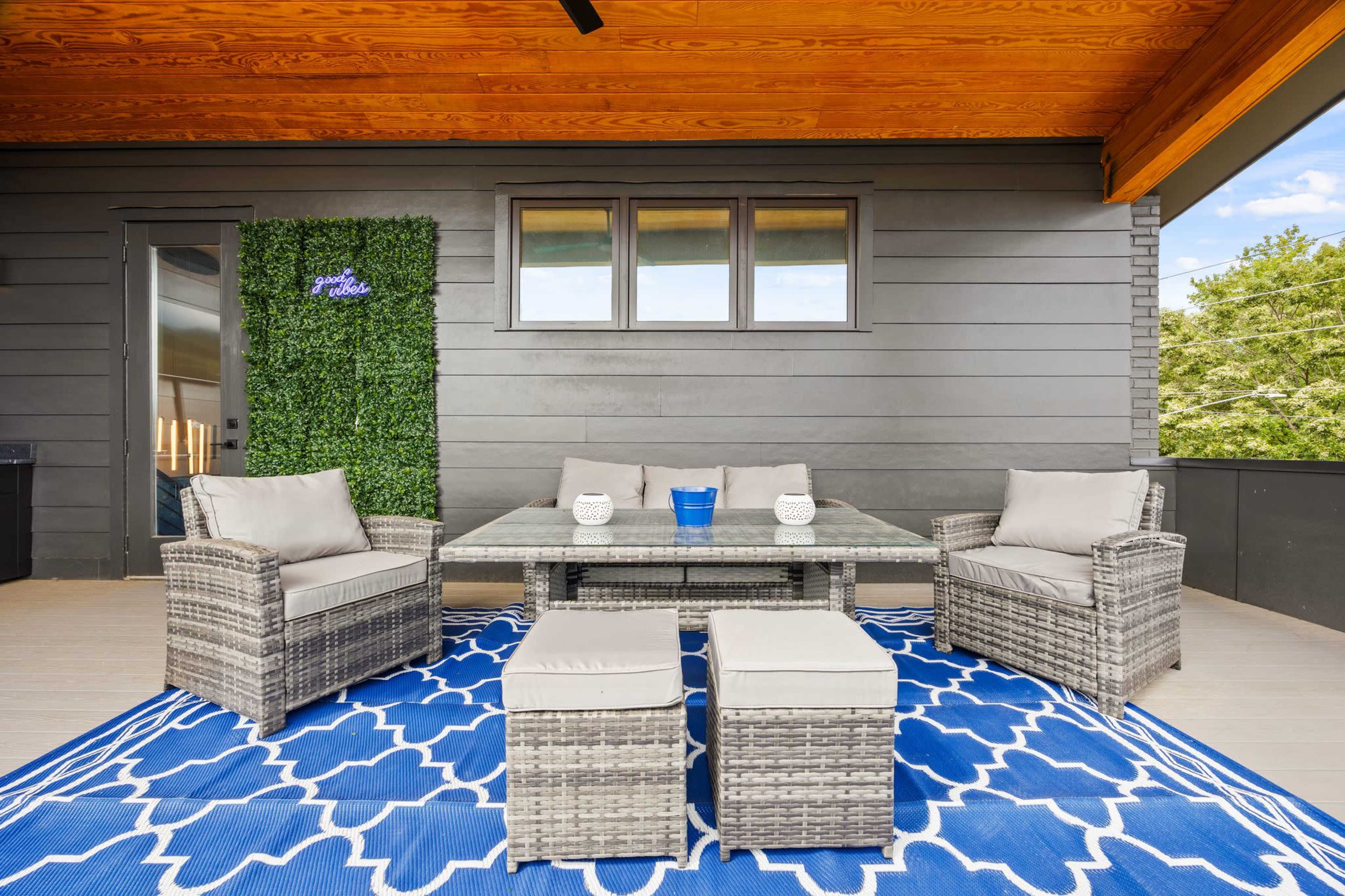 A patio area features a glass-top table surrounded by wicker chairs and ottomans on a decorative blue rug under a wooden overhang.