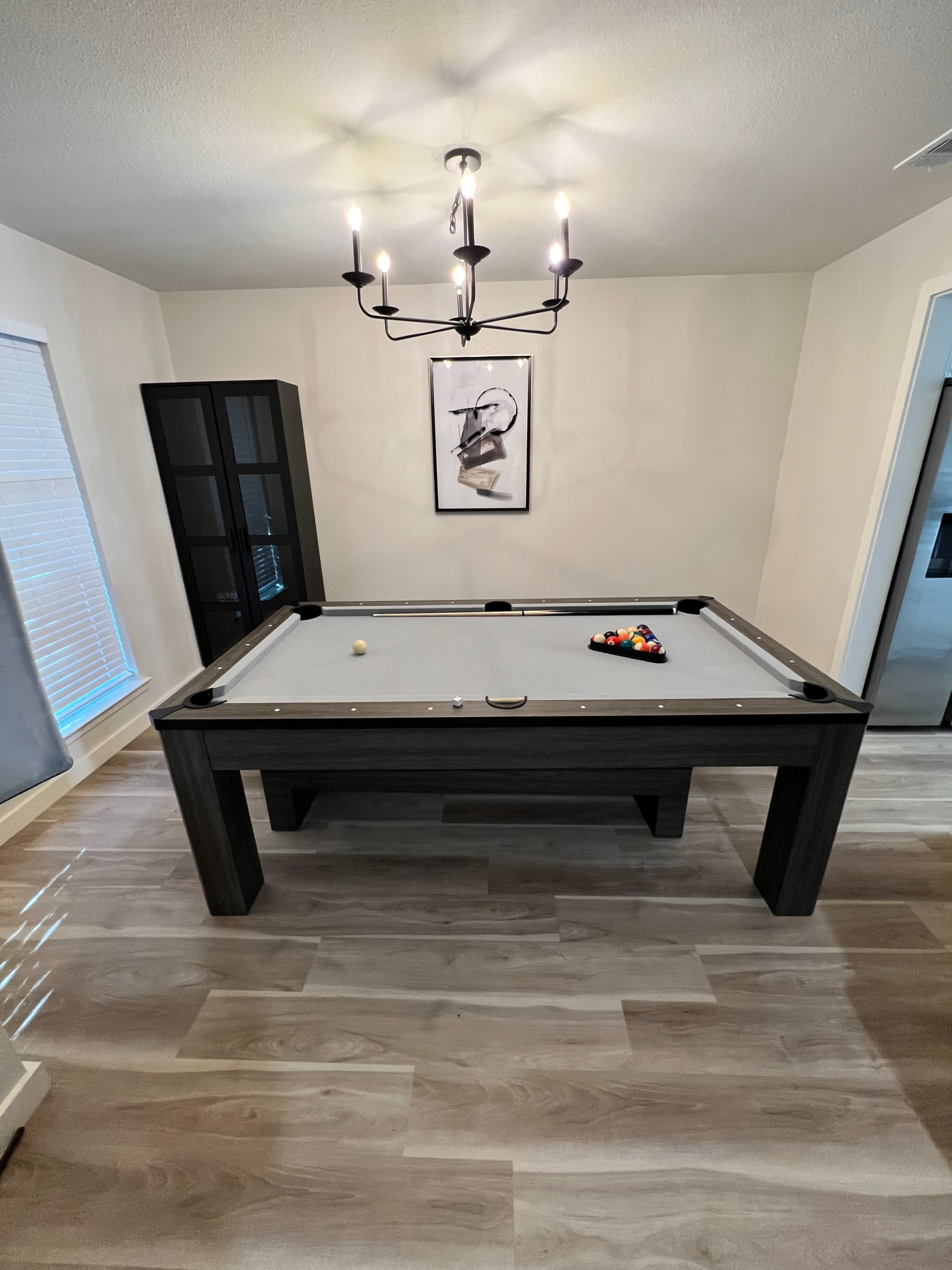 A modern billiards table with a chandelier above it, set in a room that features a large window and a tall cabinet.